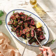 Oval platter of roasted beet chunks garnished with chopped herbs and lemon zest, served with a lemon wedge, two serving tongs, and a hand holding a fork on a rustic white table. A bottle of oil and a peach napkin are nearby.