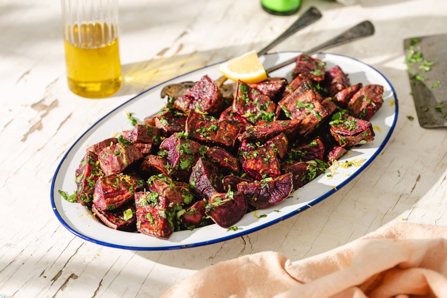 A white oval plate with roasted beetroot pieces garnished with chopped herbs and a lemon wedge, set on a rustic white table with a glass of olive oil in the background.