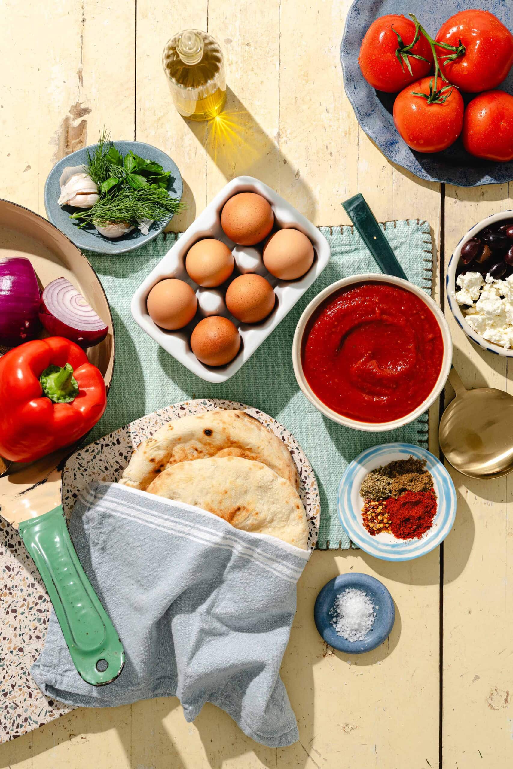 A top view of various ingredients on a table, including eggs, tomatoes, pita bread, red sauce, red onion, bell pepper, fresh herbs, oil, spices, olives, and feta cheese.
