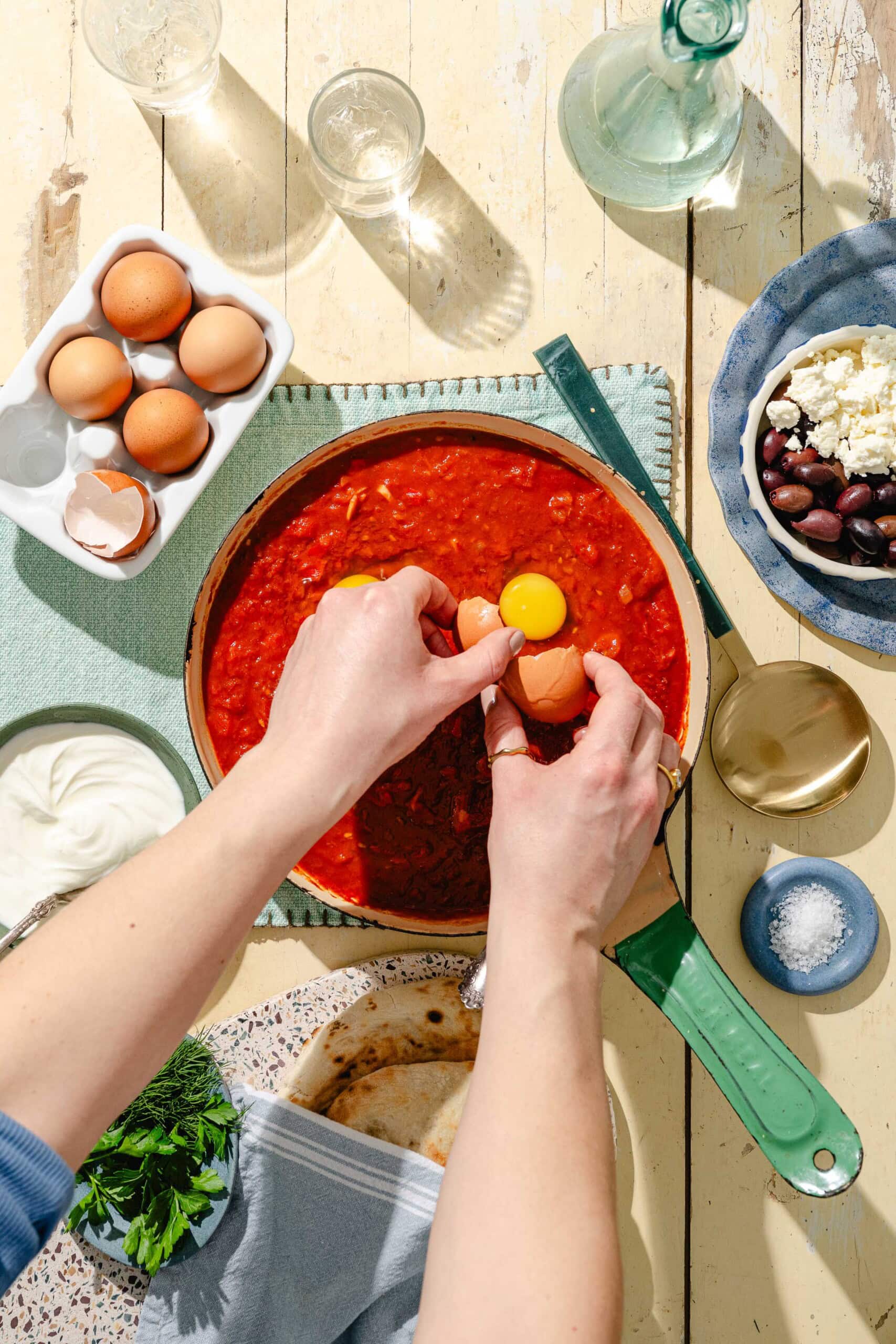 Overhead view of hands cracking an egg into a skillet of tomato sauce on a light wooden table, surrounded by ingredients like eggs, yogurt, herbs, olives, and feta cheese.