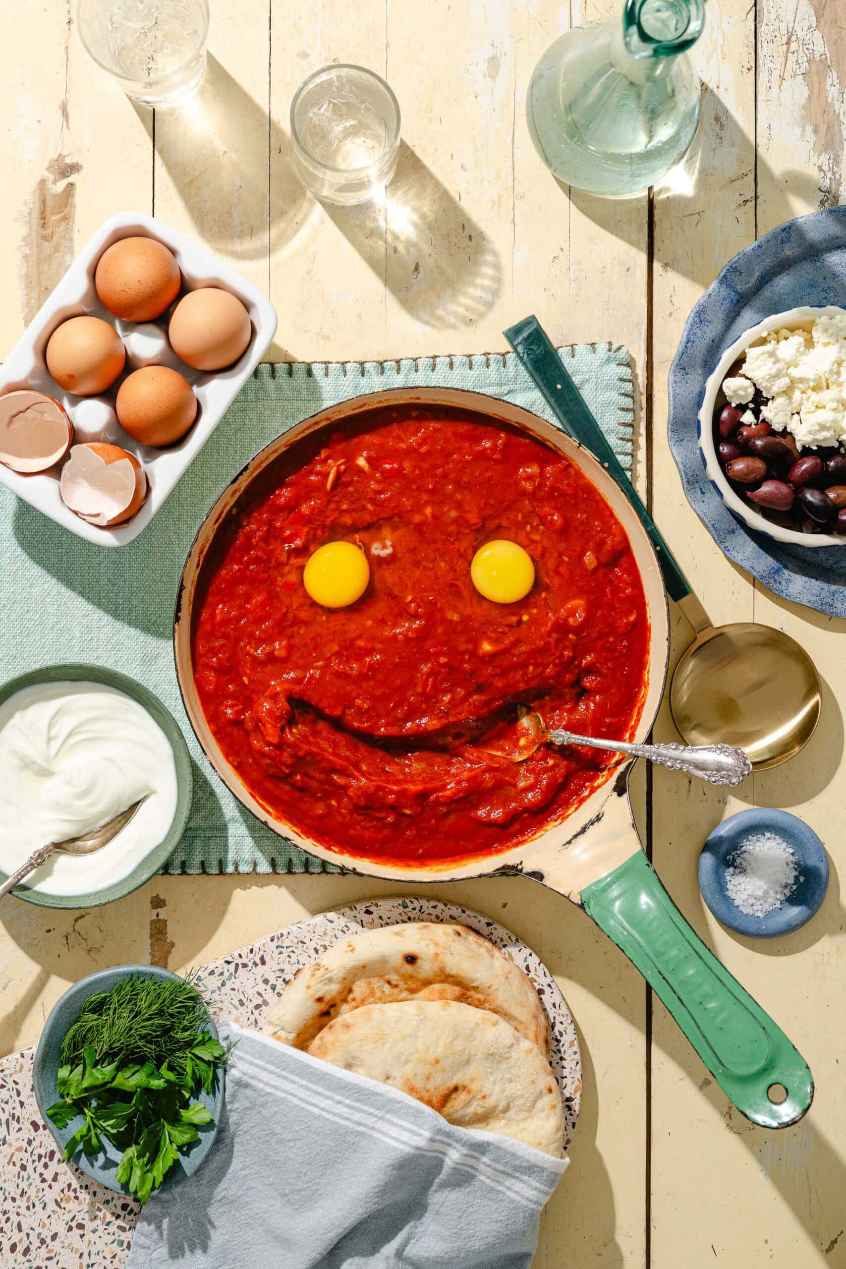 A skillet of tomato sauce with two whole eggs and a curved line creating a smiley face, surrounded by eggs, yogurt, herbs, pita bread, olives, feta cheese, and tableware on a light table.