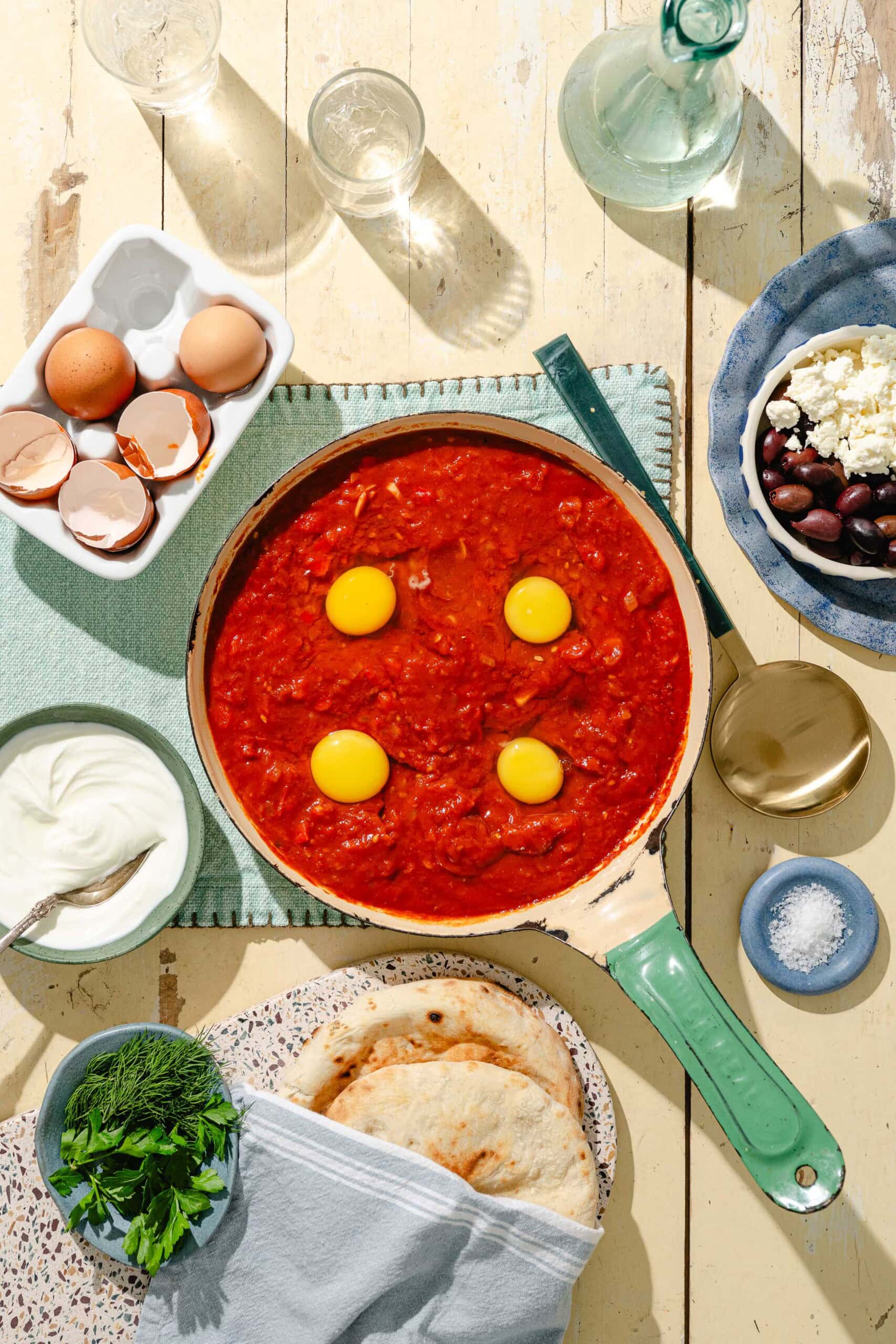 A skillet of tomato sauce with four raw eggs on top sits on a table with pita bread, fresh herbs, a bowl of olives and feta, eggshells, salt, yogurt, and glassware.