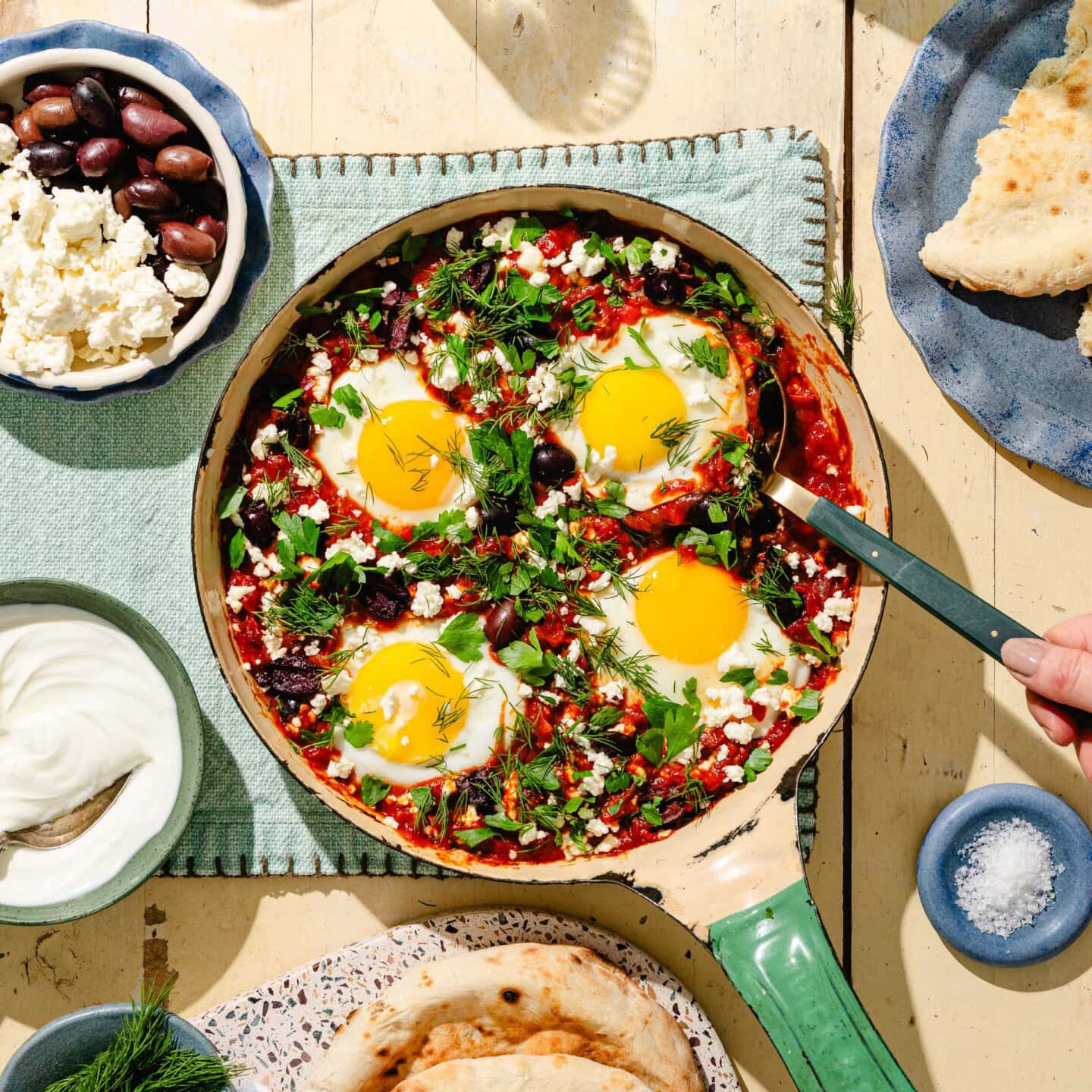 A skillet of shakshuka with eggs, tomatoes, olives, herbs, and feta cheese is surrounded by bowls of olives, feta, yogurt, pita bread, and flatbread on a wooden table.