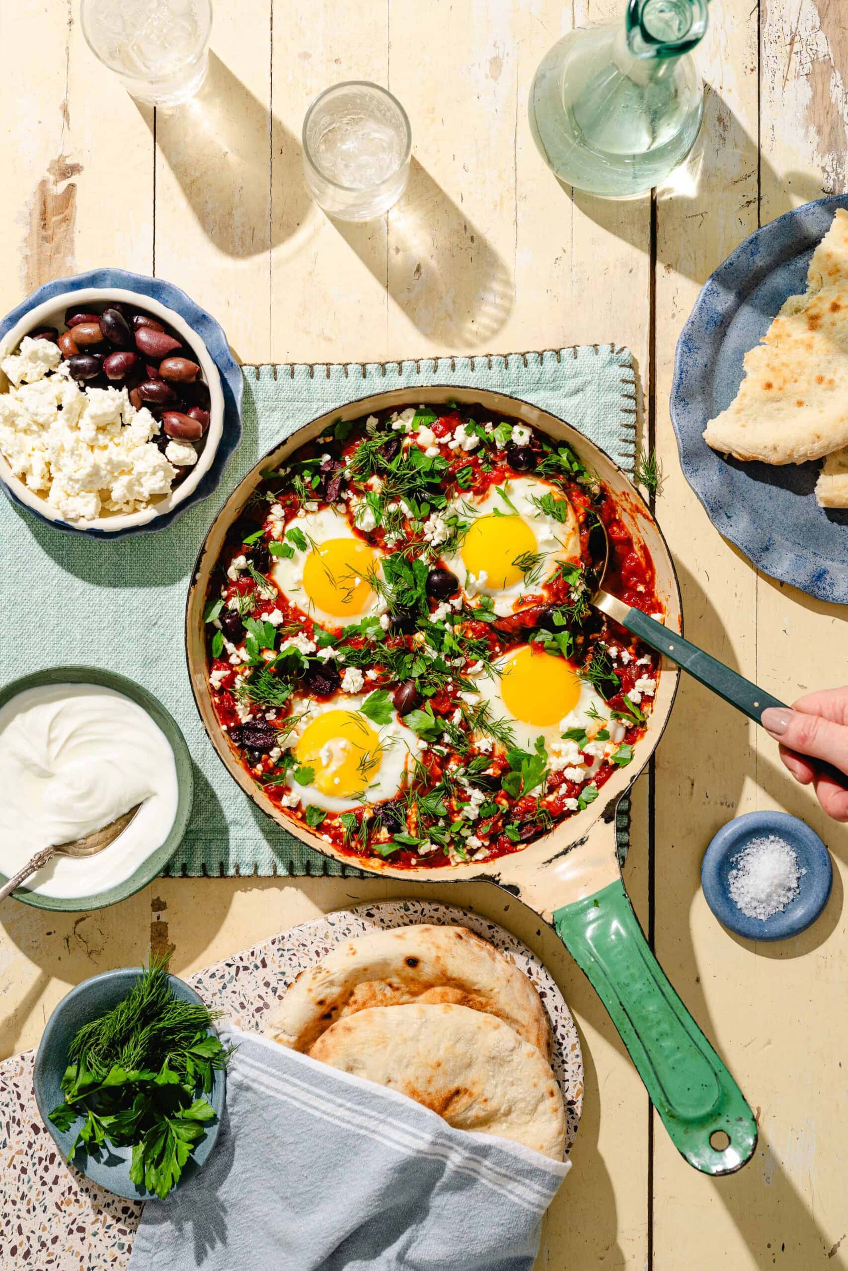 A skillet with shakshuka topped with eggs, herbs, and cheese is on a table with pita bread, a bowl of yogurt, olives, feta, fresh herbs, a bowl of salt, water glasses, and a bottle. A hand holds a fork in the pan.