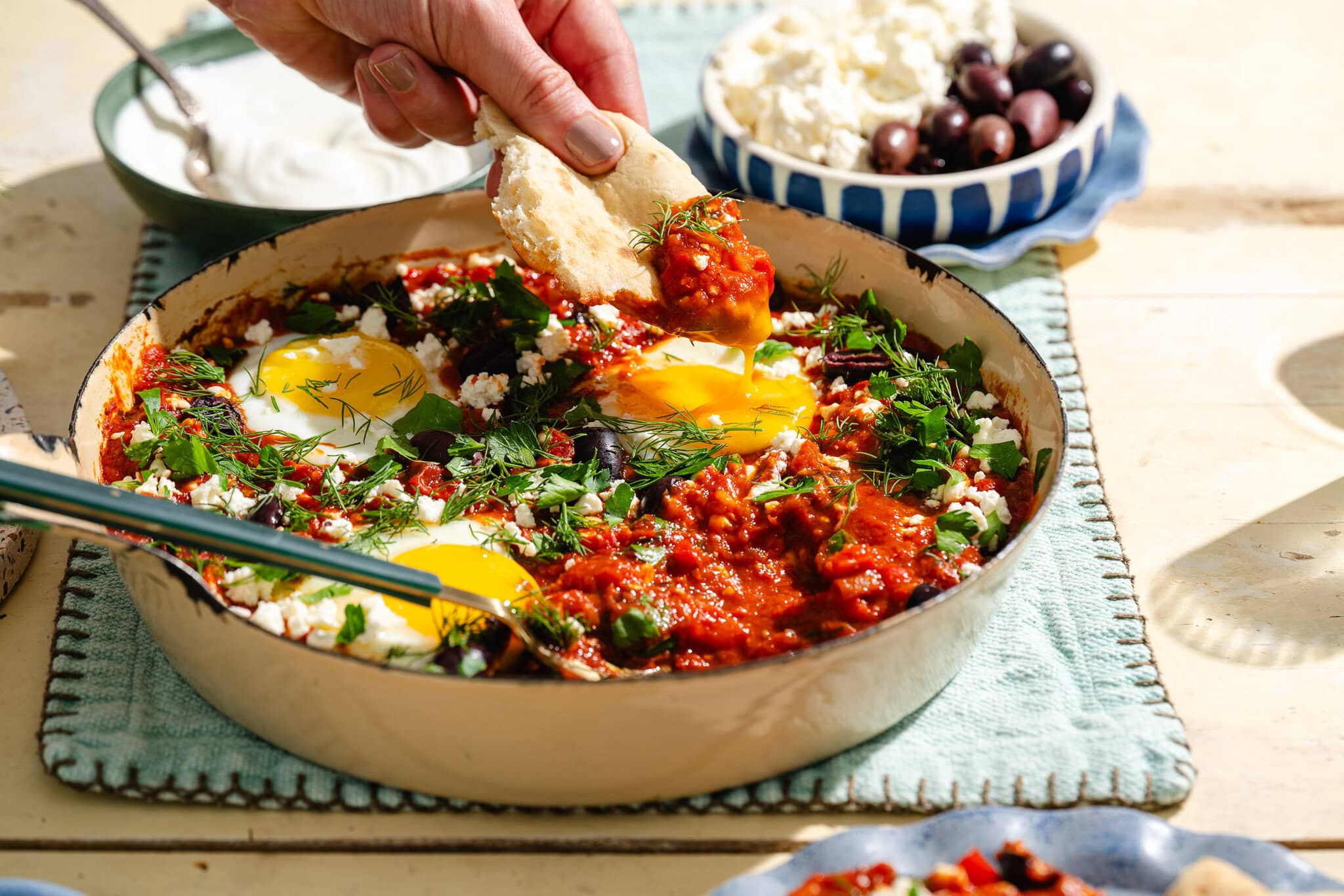 A hand dips bread into a skillet of shakshuka with poached eggs, tomato sauce, herbs, and feta. Bowls of olives and cheese sit nearby on a sunlit table.