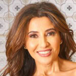 A woman with long, wavy brown hair smiles at the camera. She has subtle makeup and is standing in front of a patterned tile background.