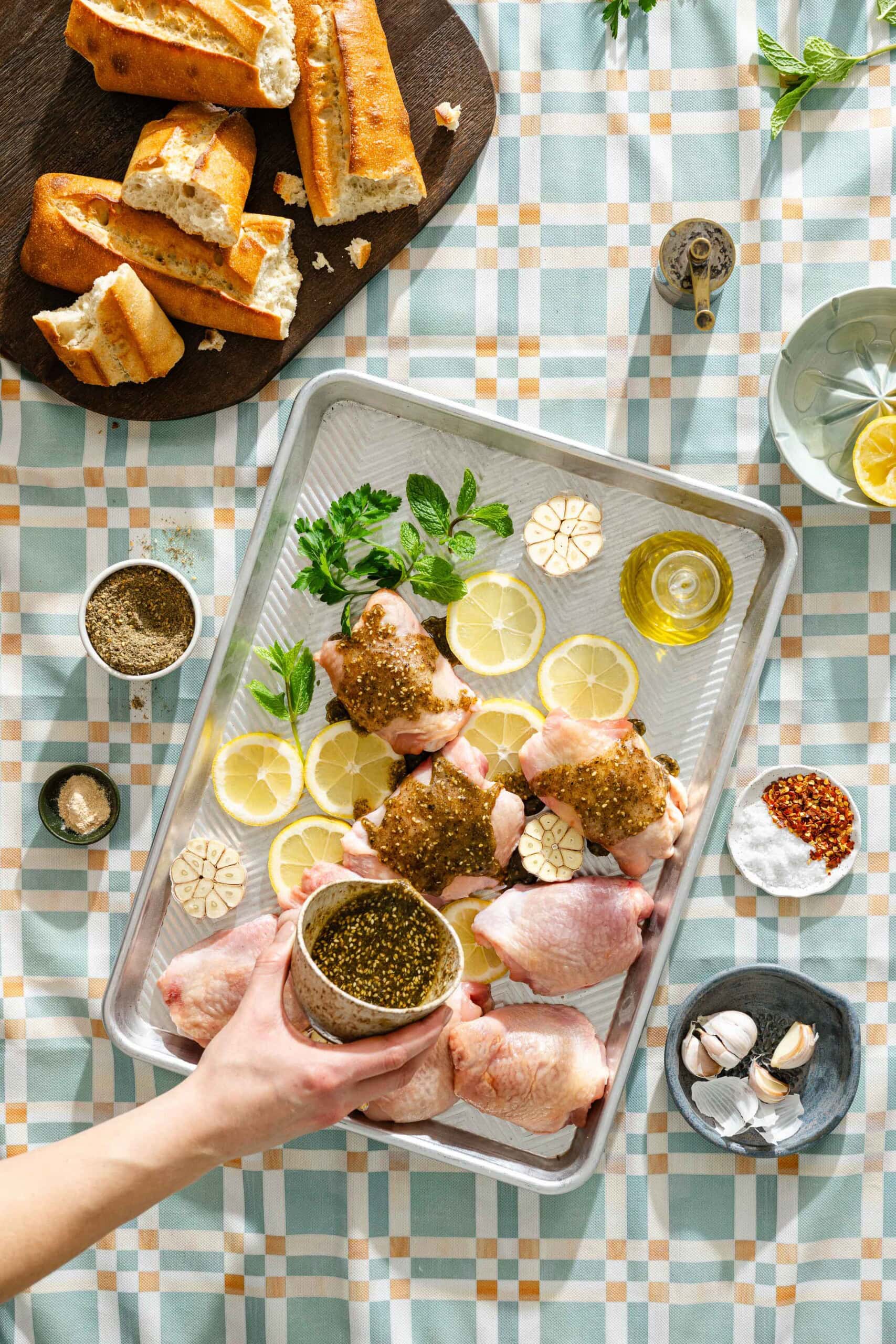 A baking tray with raw chicken thighs, lemon slices, garlic, fresh herbs, and oil sits on a patterned tablecloth; a hand sprinkles seasoning over the chicken. Next to it are bread, spices, garlic, and lemon slices.