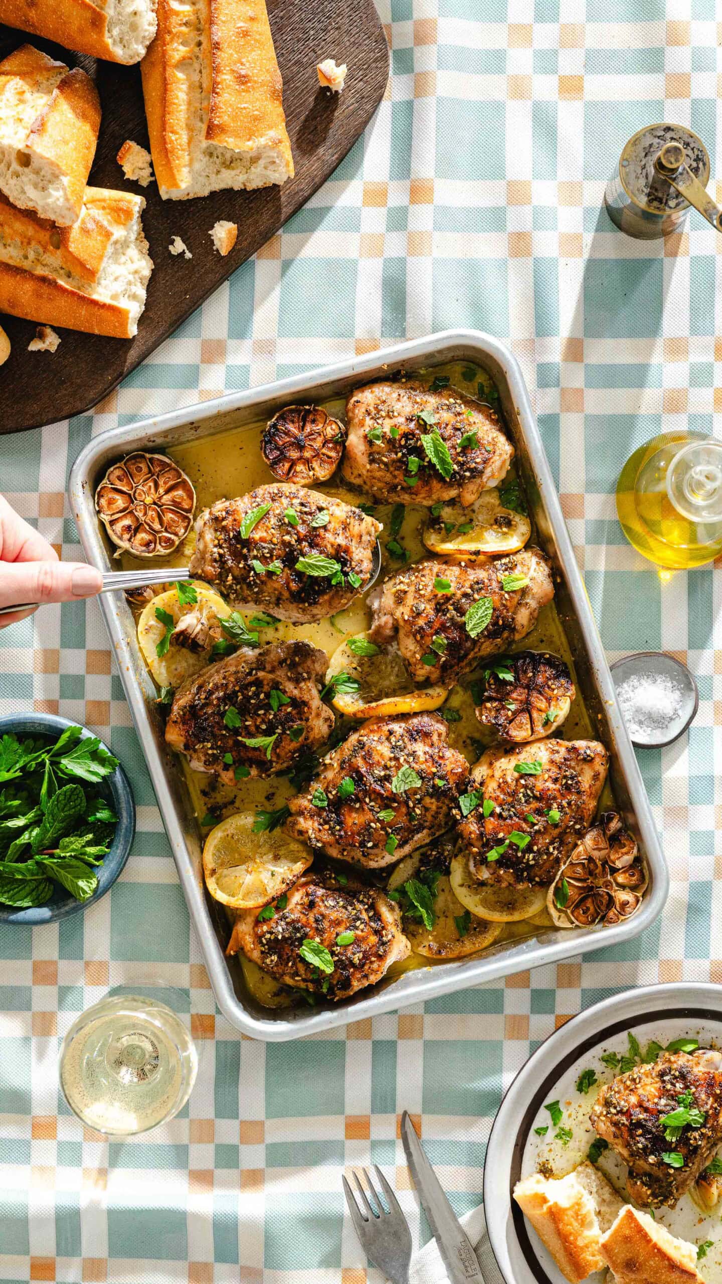 A tray of roasted chicken thighs with lemon slices and herbs sits on a checkered tablecloth. Surrounding it are bread, fresh mint, olive oil, a glass of white wine, and utensils, with a hand reaching for a piece.