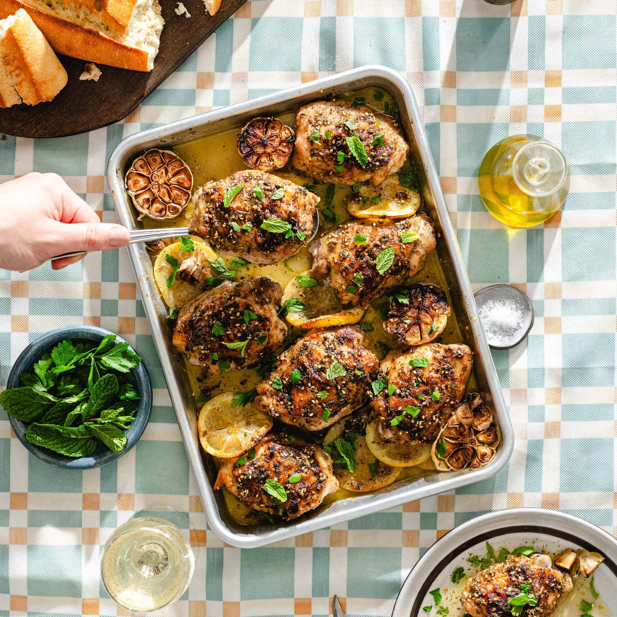 A hand serving roasted chicken thighs garnished with herbs, lemon slices, and roasted garlic from a baking dish. The table is set with bread, olive oil, mint leaves, and wine on a pastel checkered tablecloth.
