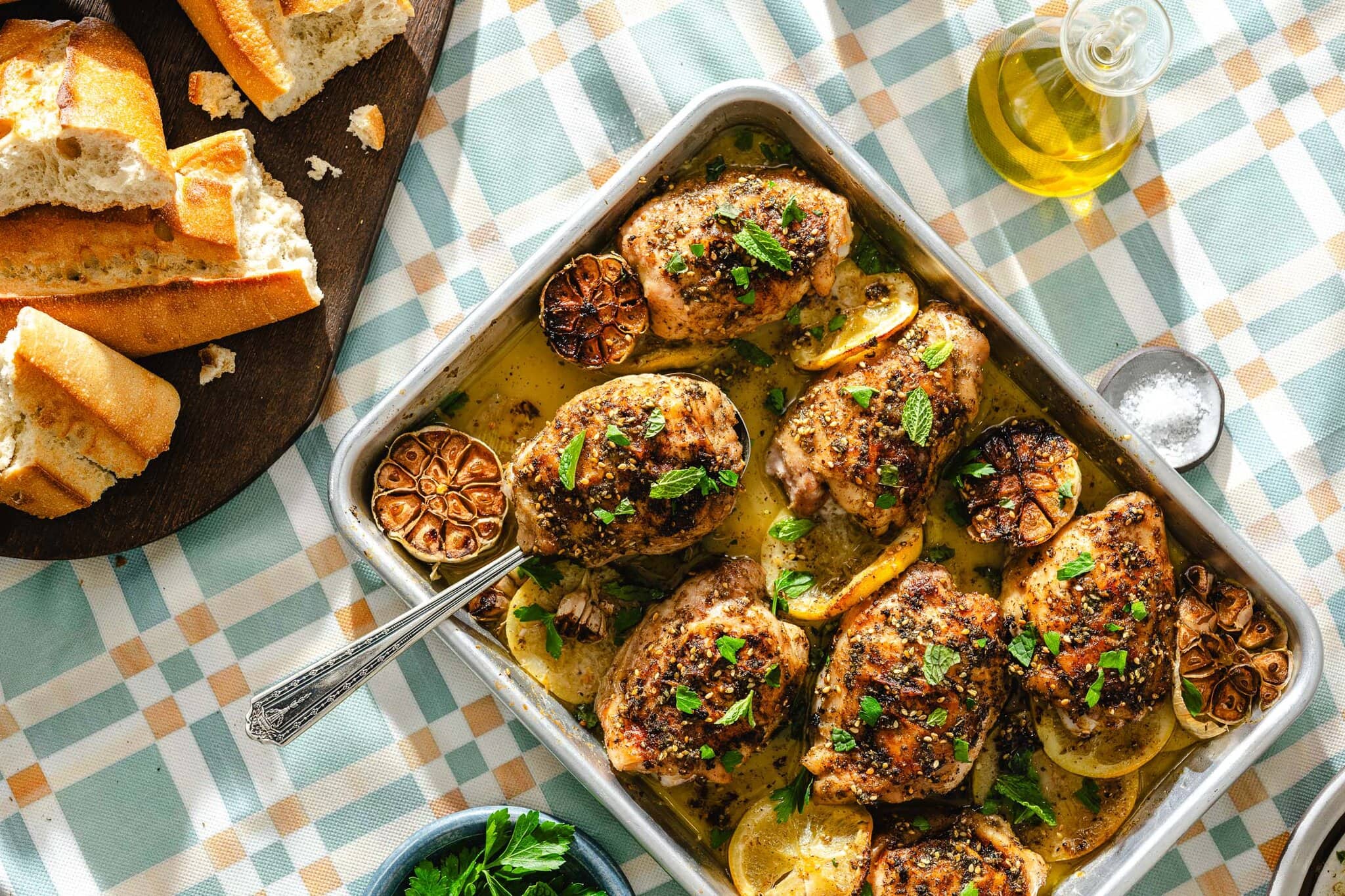 Baked chicken thighs with herbs and lemon slices in a tray, garnished with fresh herbs and roasted garlic, served with sliced bread and a bottle of olive oil on a checkered tablecloth.