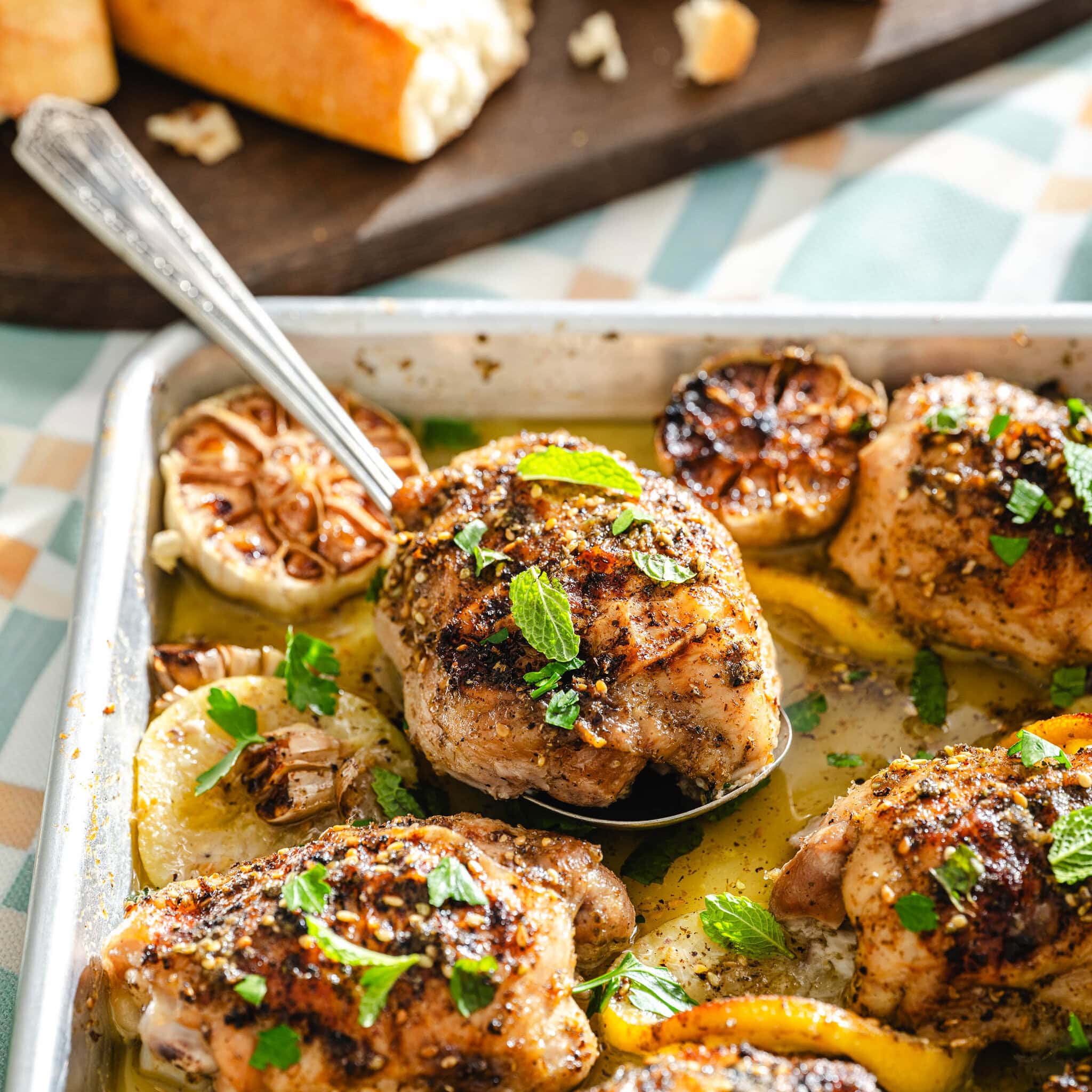 A baking tray with roasted chicken thighs, lemon slices, and halved garlic heads, garnished with fresh herbs. A spoon lifts one piece, and bread is visible on a board in the background.