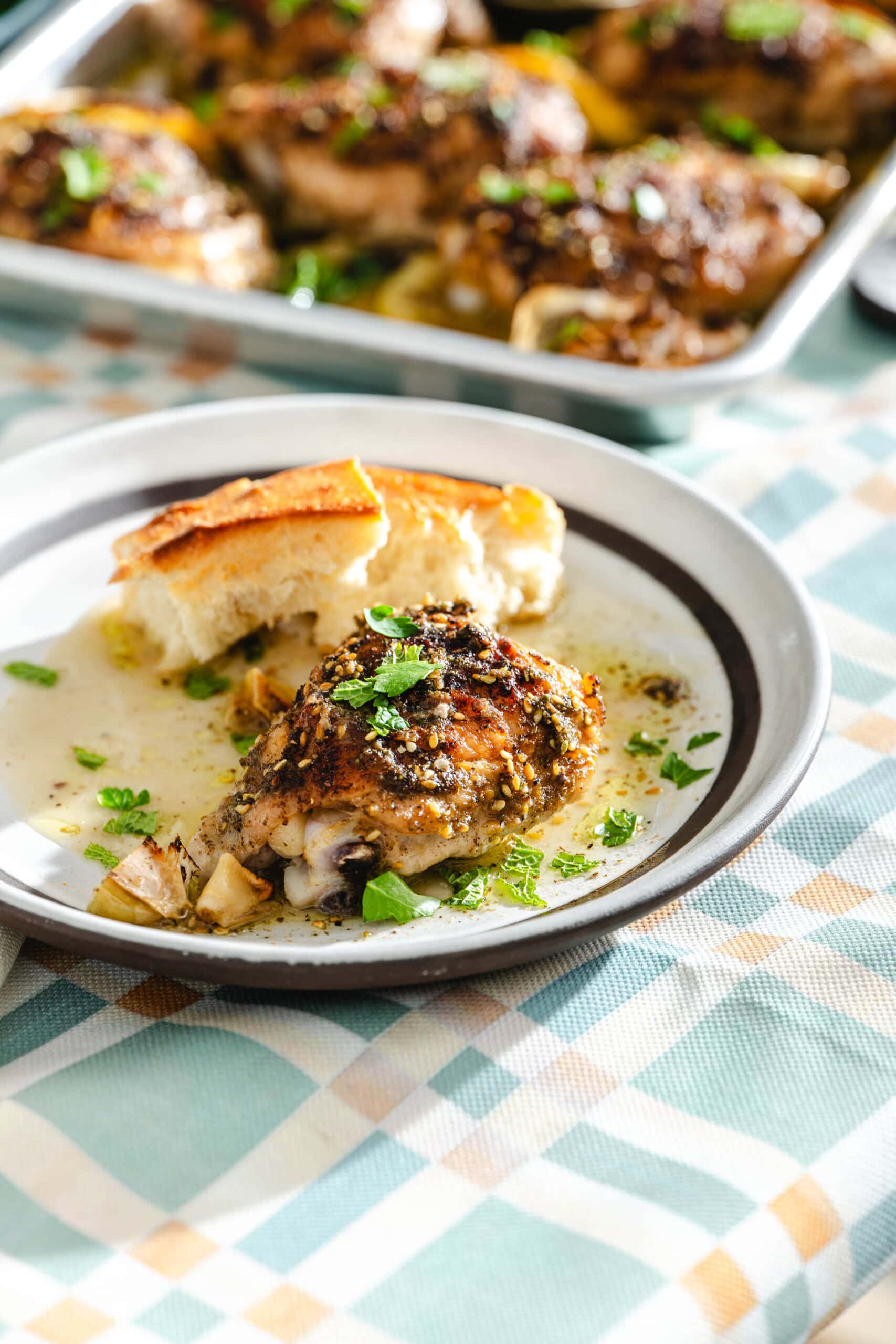A plate with a seasoned baked chicken thigh, garnished with herbs and served with a slice of bread. A tray of more baked chicken is visible in the background on a checkered tablecloth.