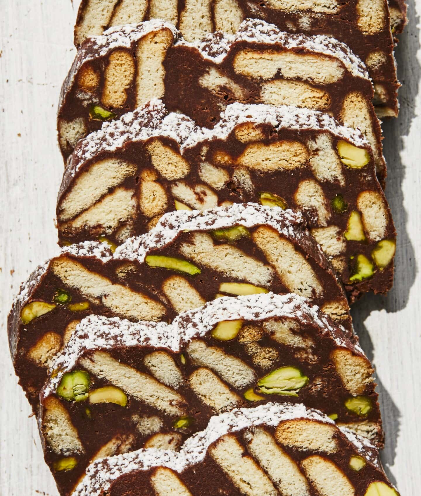 Slices of chocolate biscuit cake with visible pieces of biscuits and green pistachios, dusted lightly with powdered sugar, arranged in a row on a light-colored surface.