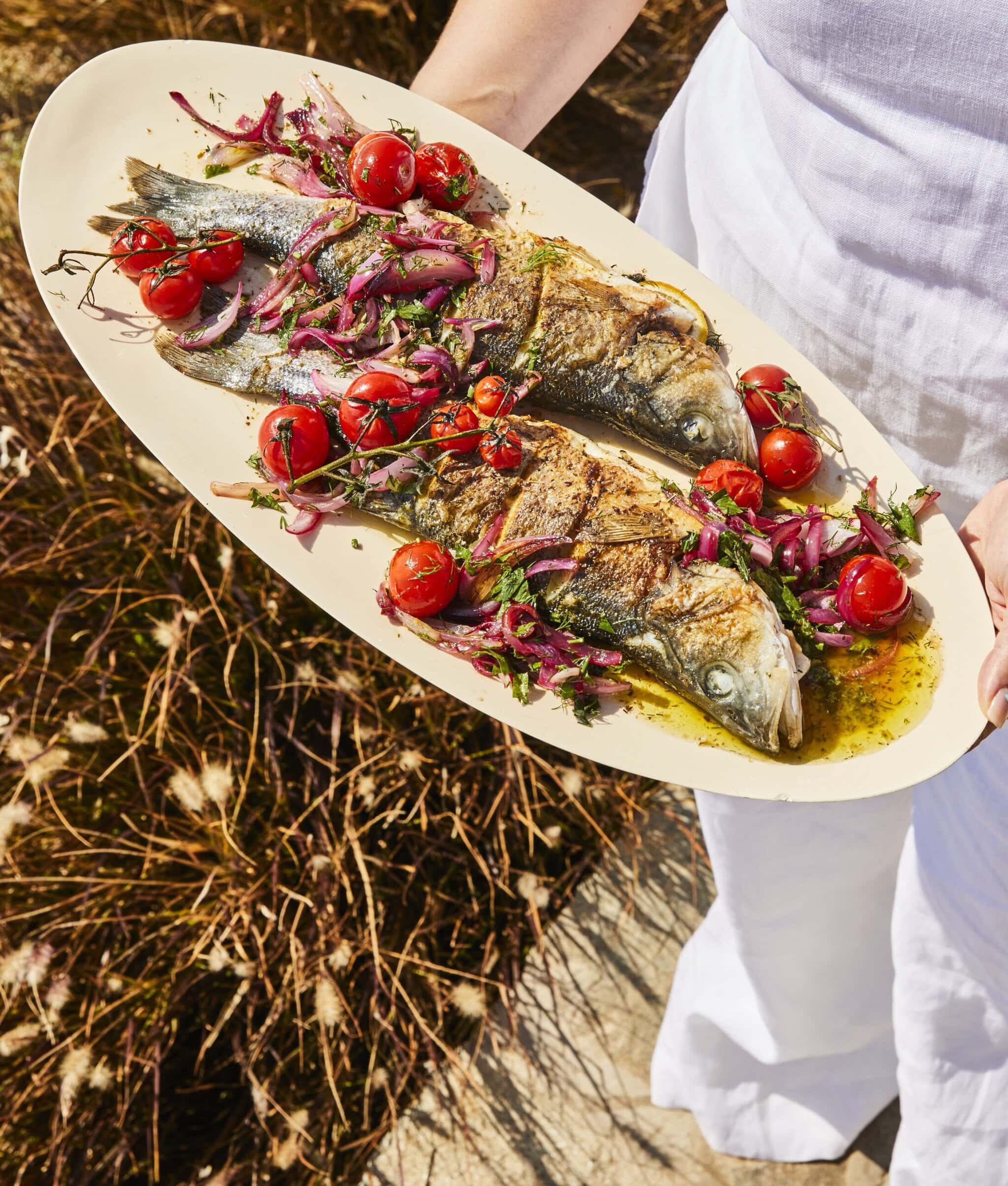 A person in white clothing holds an oval platter with two whole roasted fish, garnished with cherry tomatoes, red onions, herbs, and sauce, outdoors in sunlight.