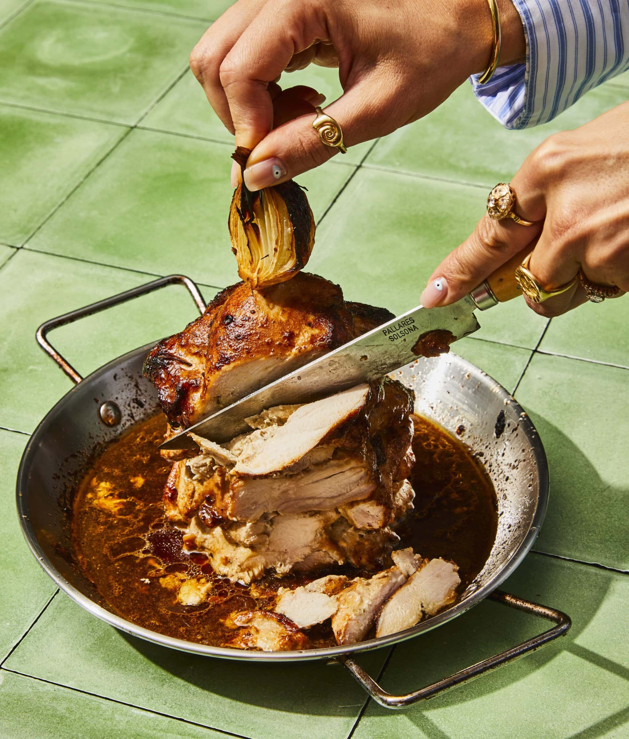 A person slices a juicy, browned roast in a pan filled with rich sauce on a green-tiled surface, holding an onion with one hand and cutting meat with a knife in the other.