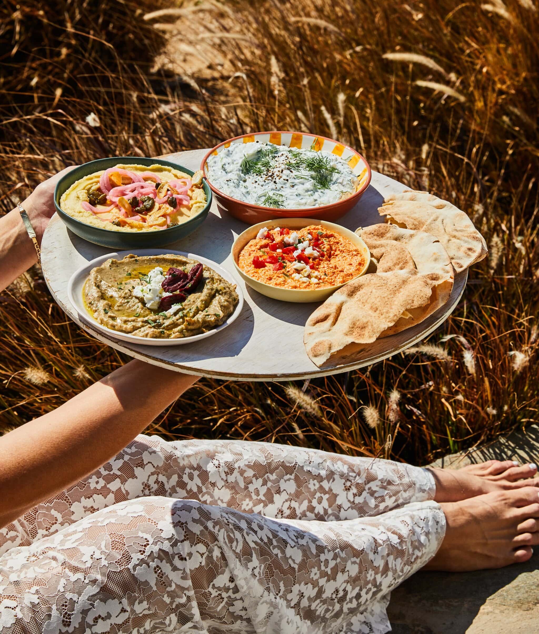 A person in a white lace dress holds a round tray with bowls of dips, including tzatziki, hummus, baba ganoush, and another dip, along with pieces of pita bread, outdoors by tall grass.