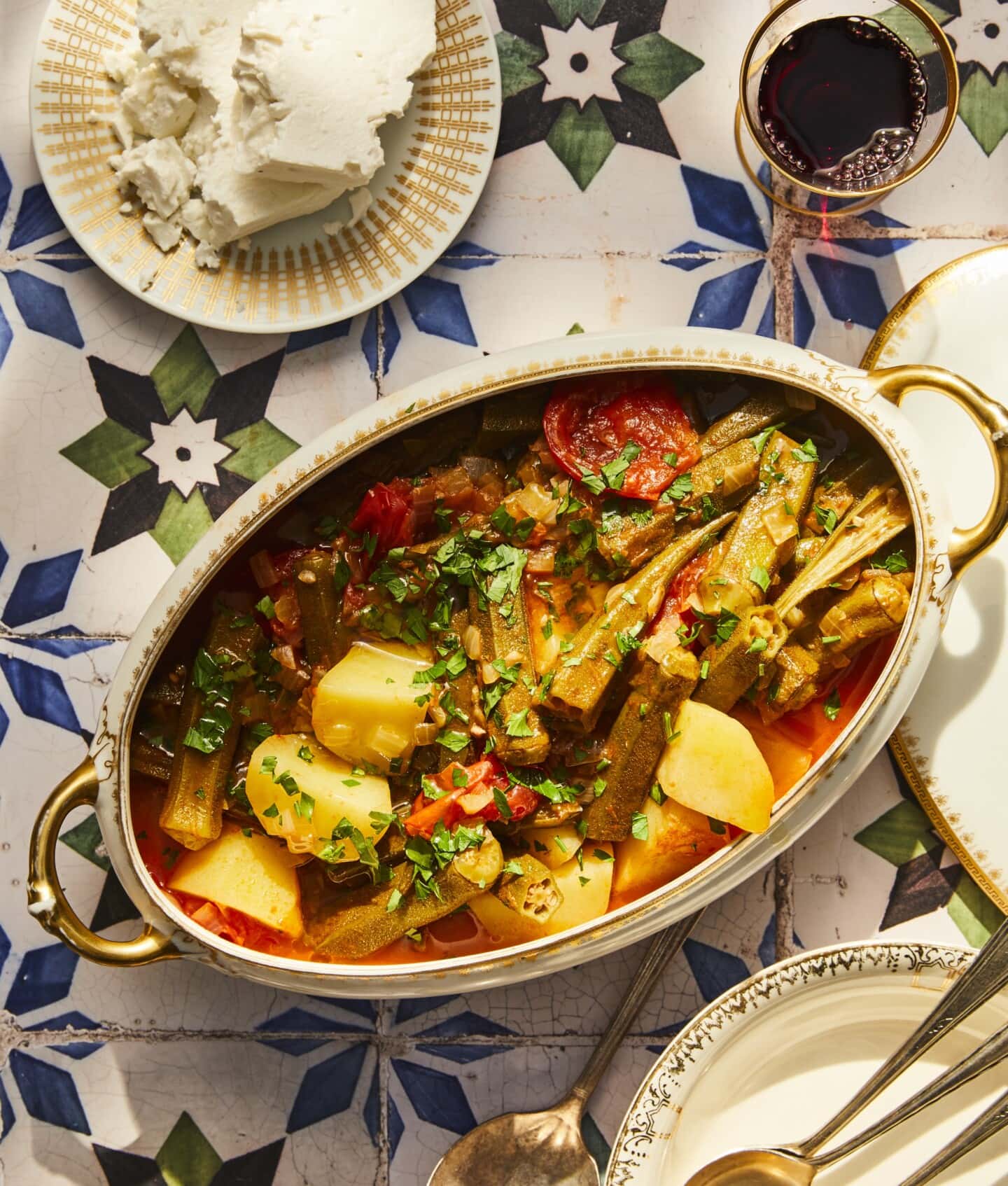 Oval dish with okra stew, potatoes, and tomatoes, garnished with chopped parsley. Nearby are a plate of soft cheese, a glass of red wine, and decorative plates and spoons on a patterned tile surface.