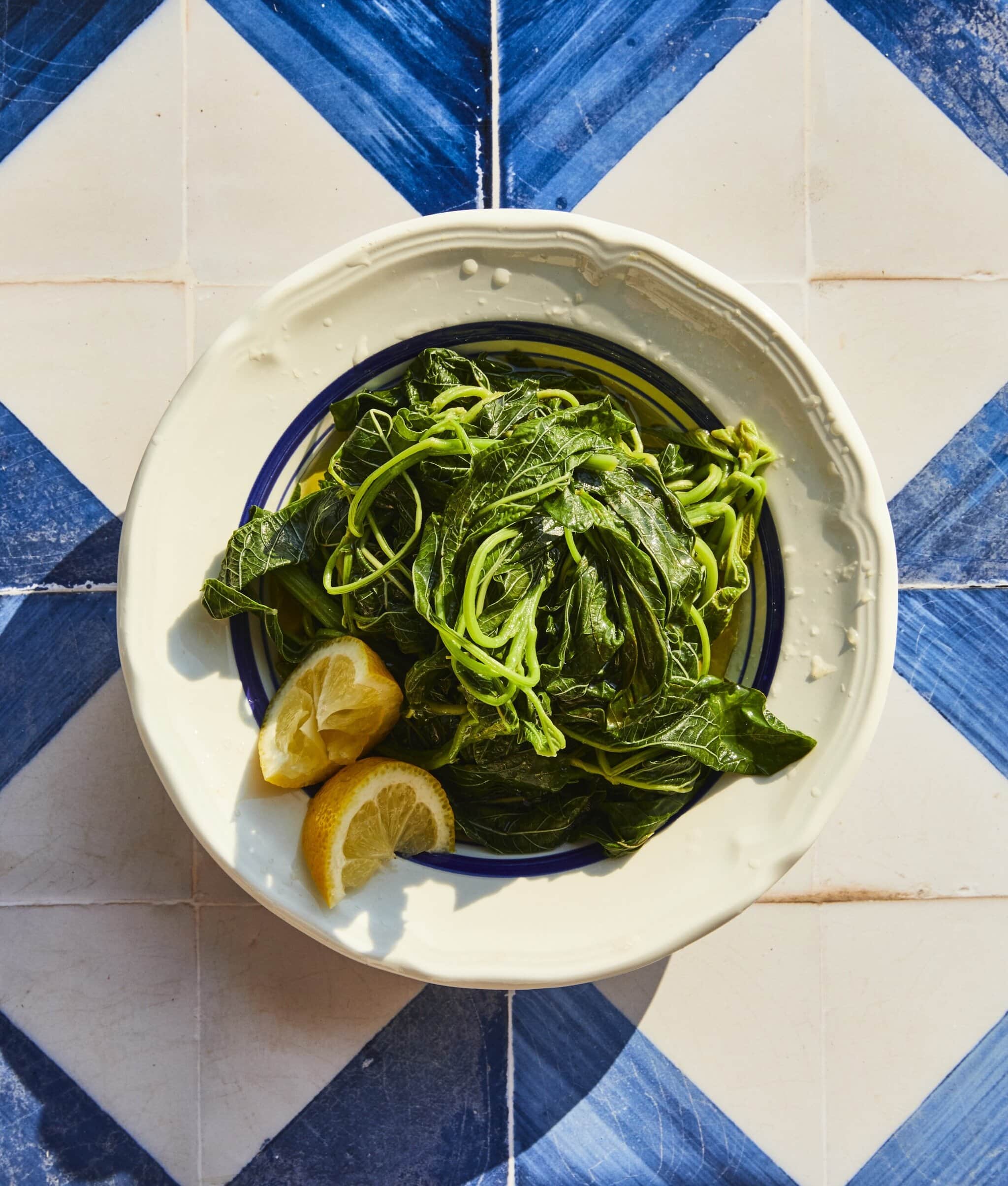 A bowl of cooked leafy greens with two lemon wedges sits on a white and blue tiled surface.
