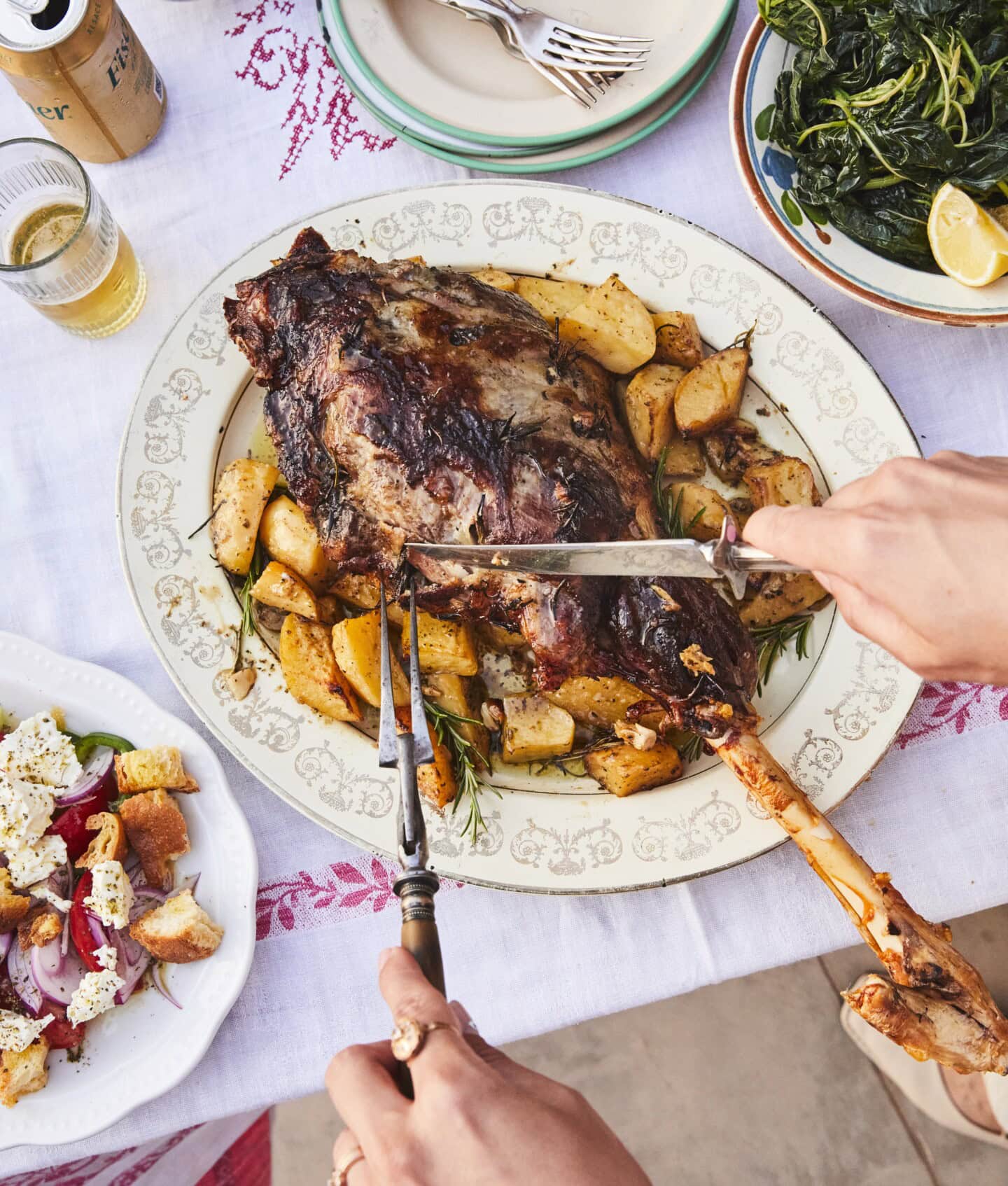 A person carves a roasted leg of lamb with potatoes on a decorative platter, surrounded by plates of cooked greens and salad on a white tablecloth.