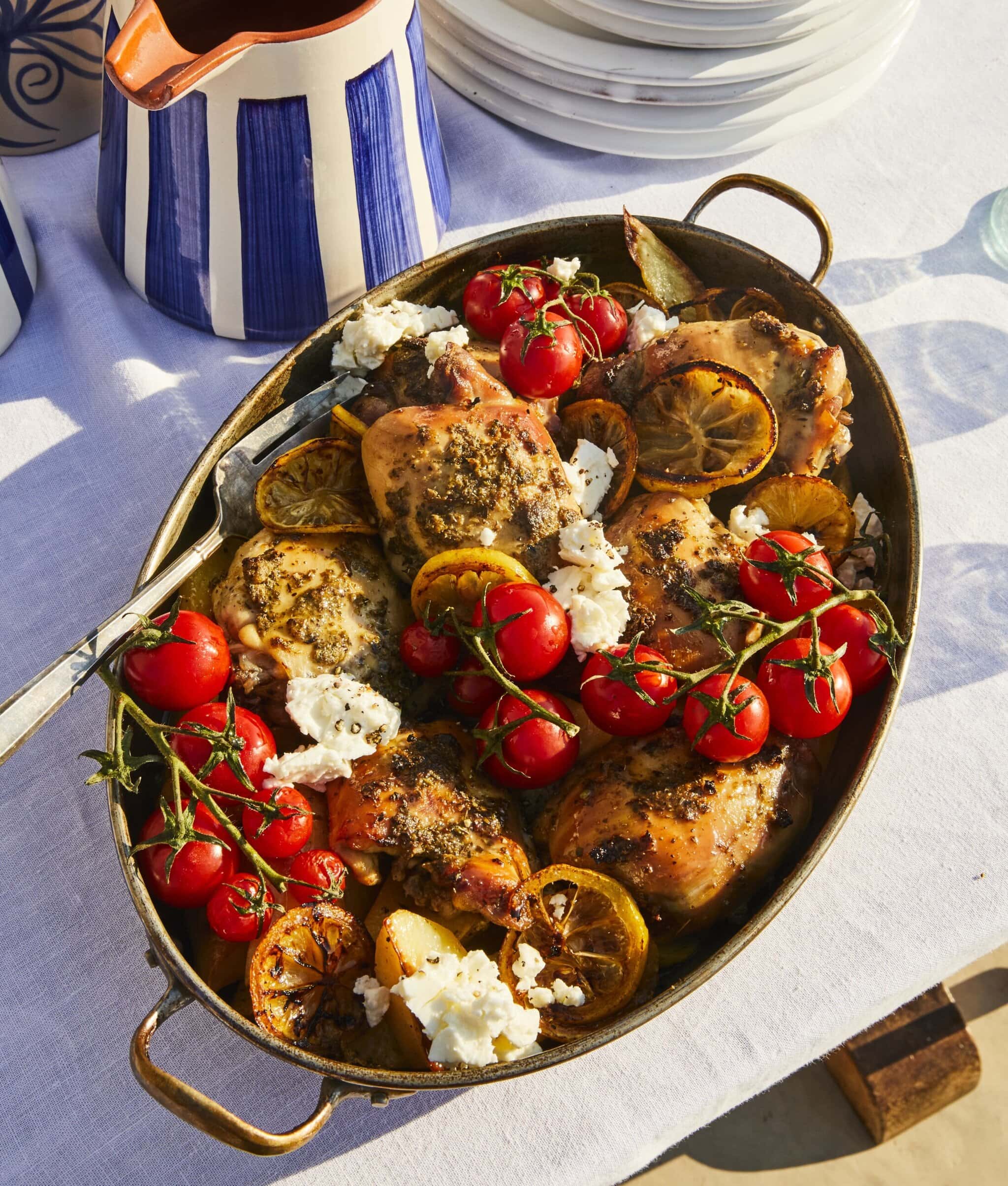 A serving dish filled with roasted chicken pieces, lemon slices, vine tomatoes, and dollops of white cheese, placed on a white tablecloth next to a blue-and-white striped pitcher and stacked white plates.