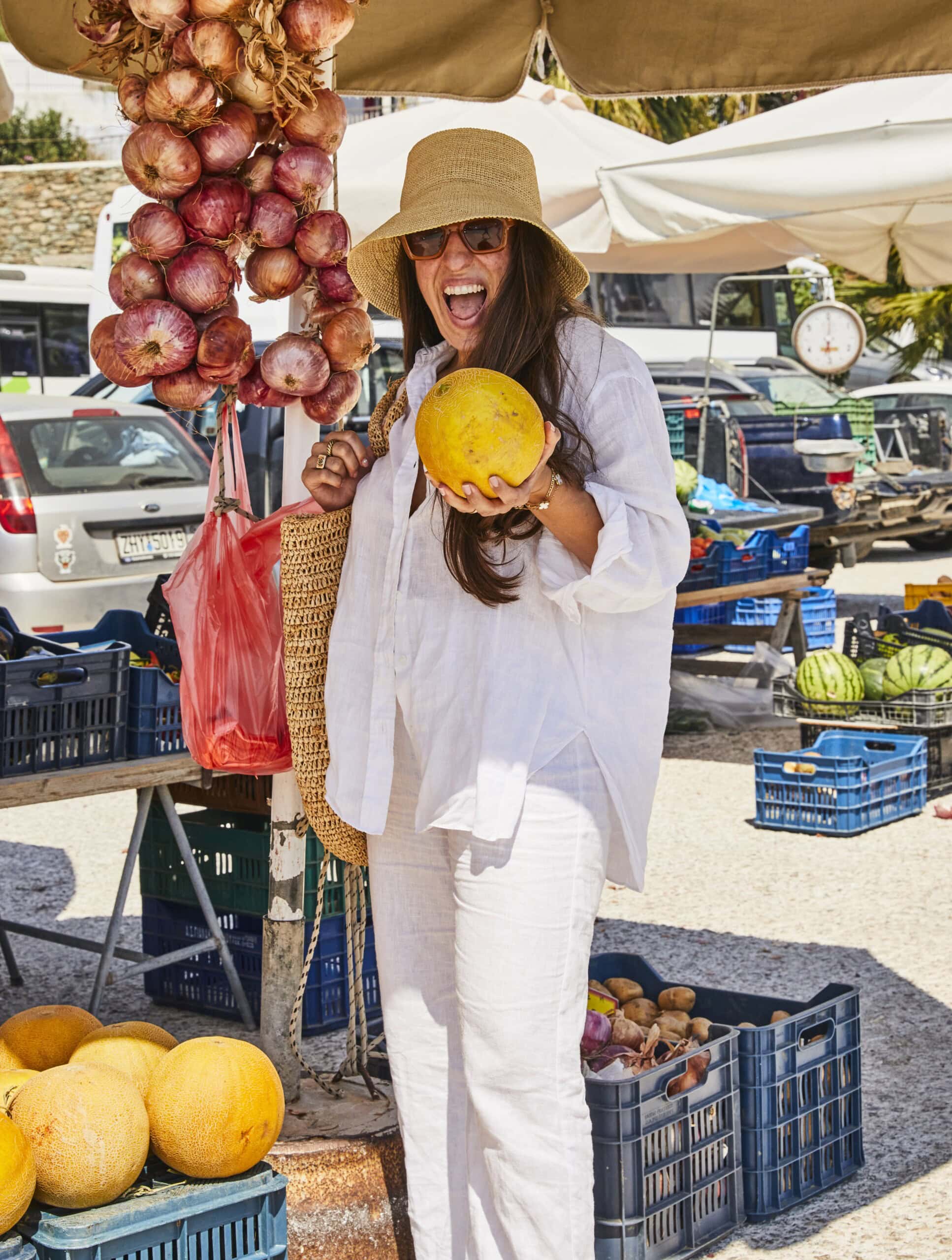 A smiling woman in a sunhat and white outfit holds a yellow melon at an outdoor market. She stands near crates of melons and hanging onions, with market stalls and vehicles visible in the background.