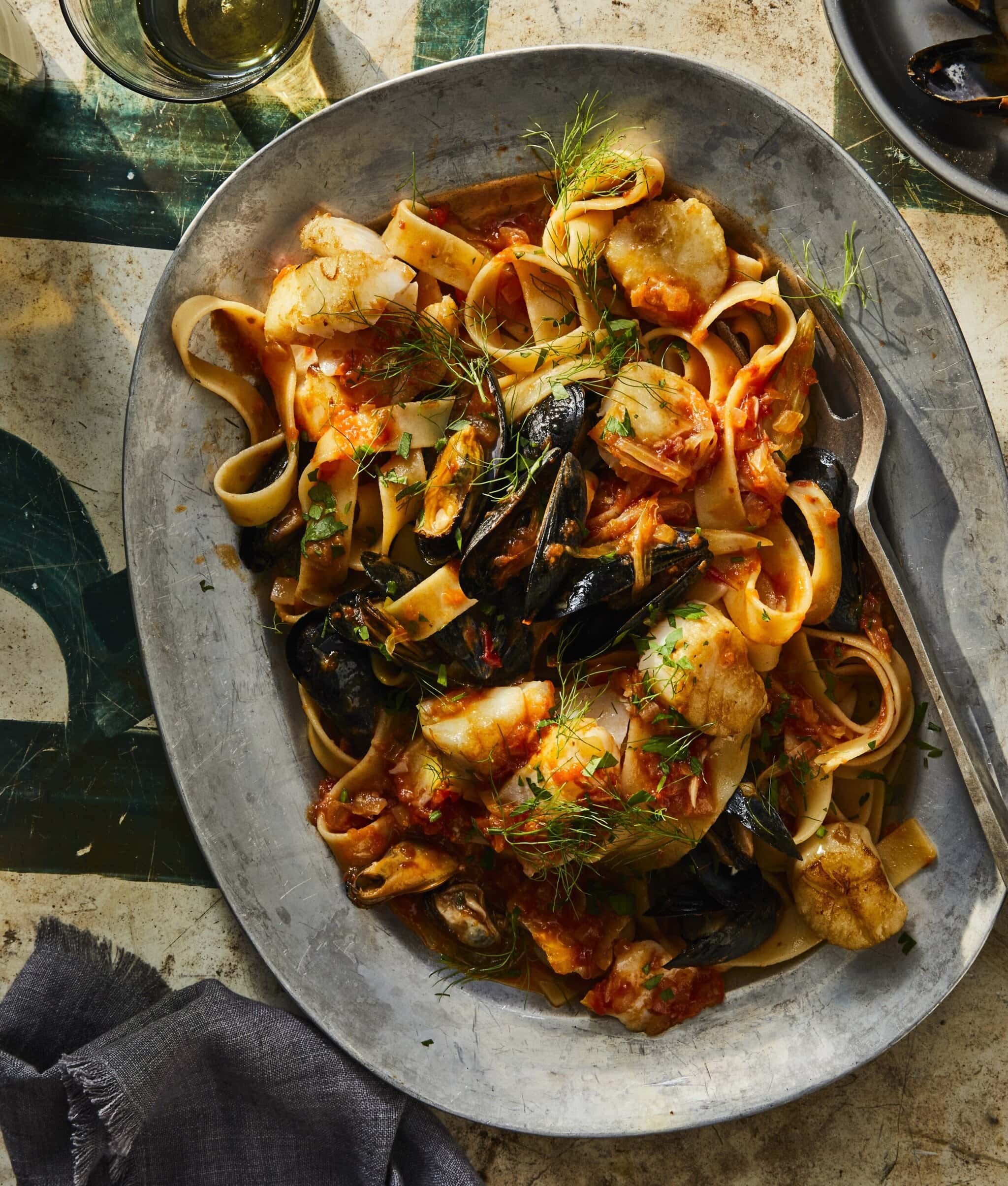 A large oval platter filled with pasta, mussels, chunks of fish, tomato sauce, and fresh herbs, sitting on a rustic table with a fork, a napkin, and a glass of white wine nearby.
