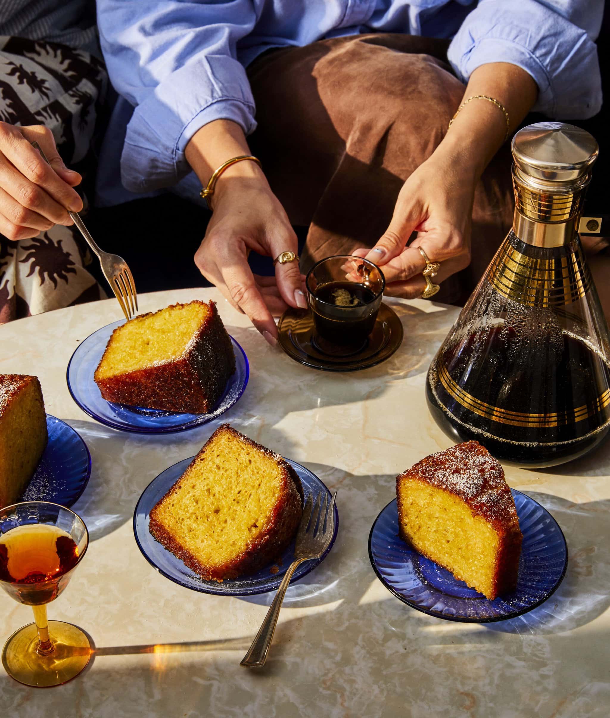 Three slices of yellow cake on blue plates, served with small cups of coffee and a glass carafe, as two people reach for their drinks at a sunlit table.