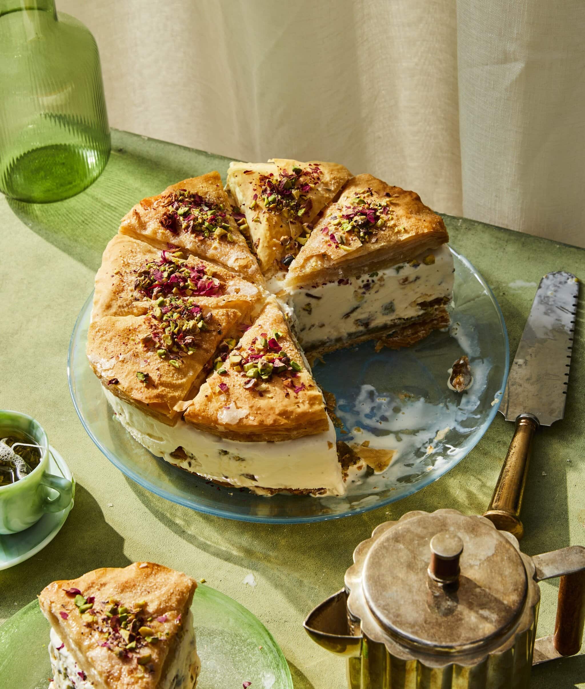A round, layered cake with creamy filling and golden pastry top, garnished with chopped pistachios and rose petals, sits on a glass plate. A slice is cut out, and a cake server and teapot are nearby on a green table.