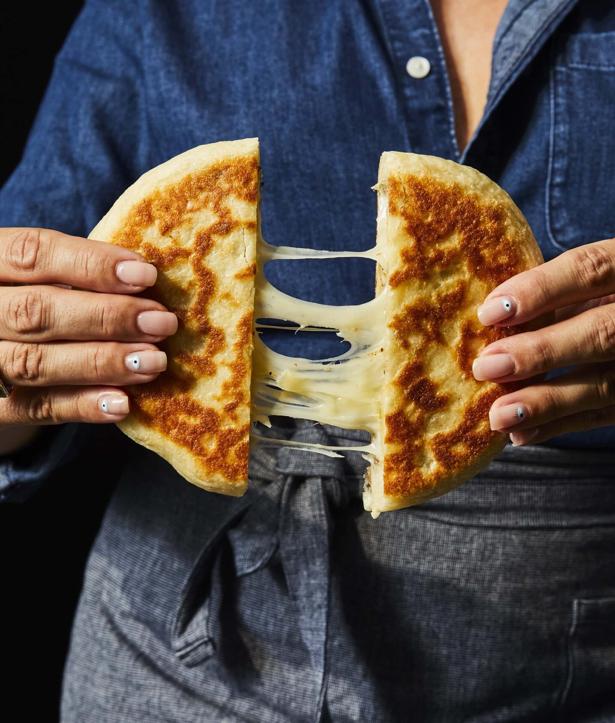 A person in a denim shirt and apron pulls apart a golden, pan-fried stuffed bread, revealing stretchy, melted cheese inside.