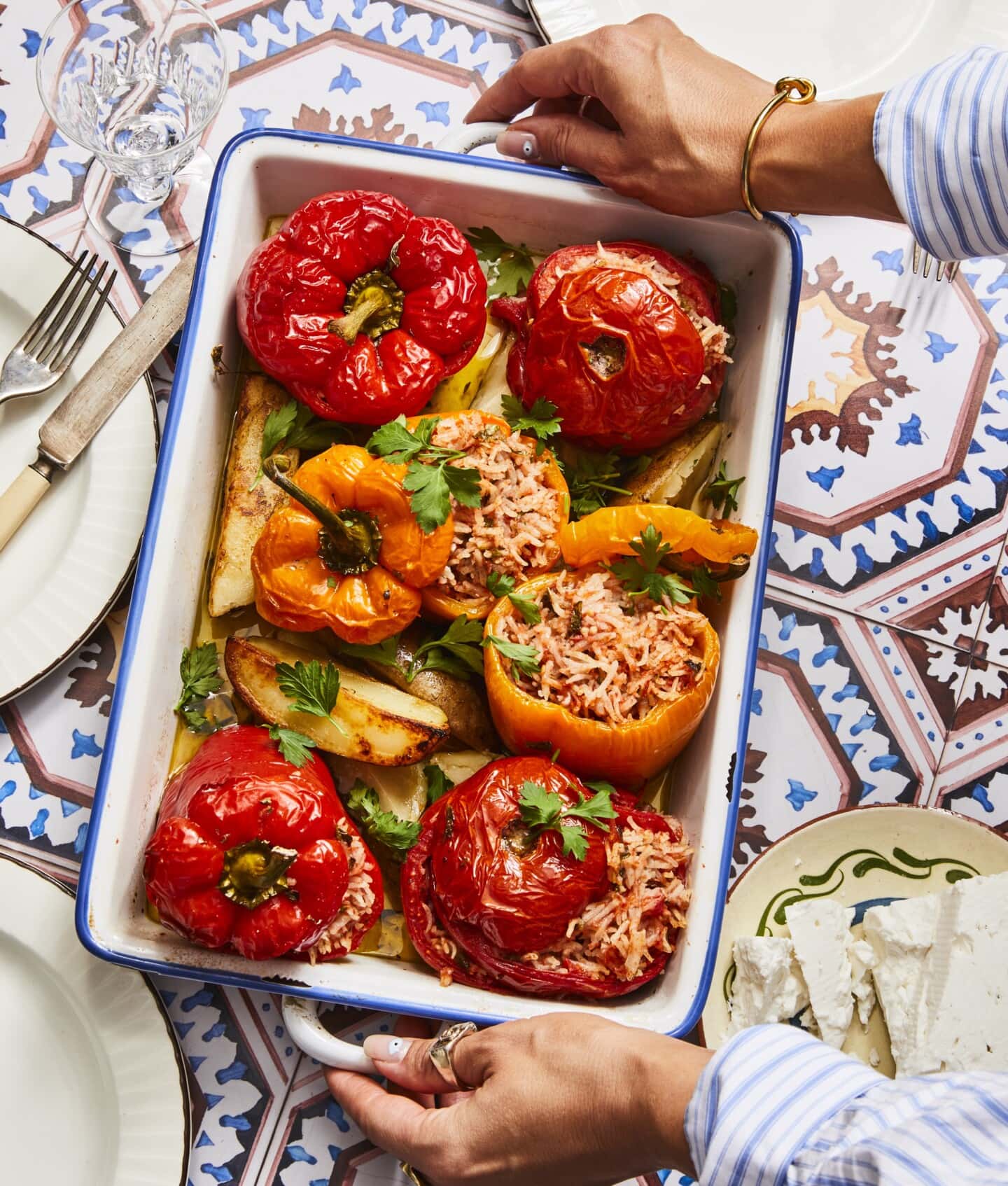 A person holds a baking dish filled with stuffed bell peppers and tomatoes atop roasted potato wedges, garnished with parsley, on a table with decorative tiles and a plate of feta cheese.
