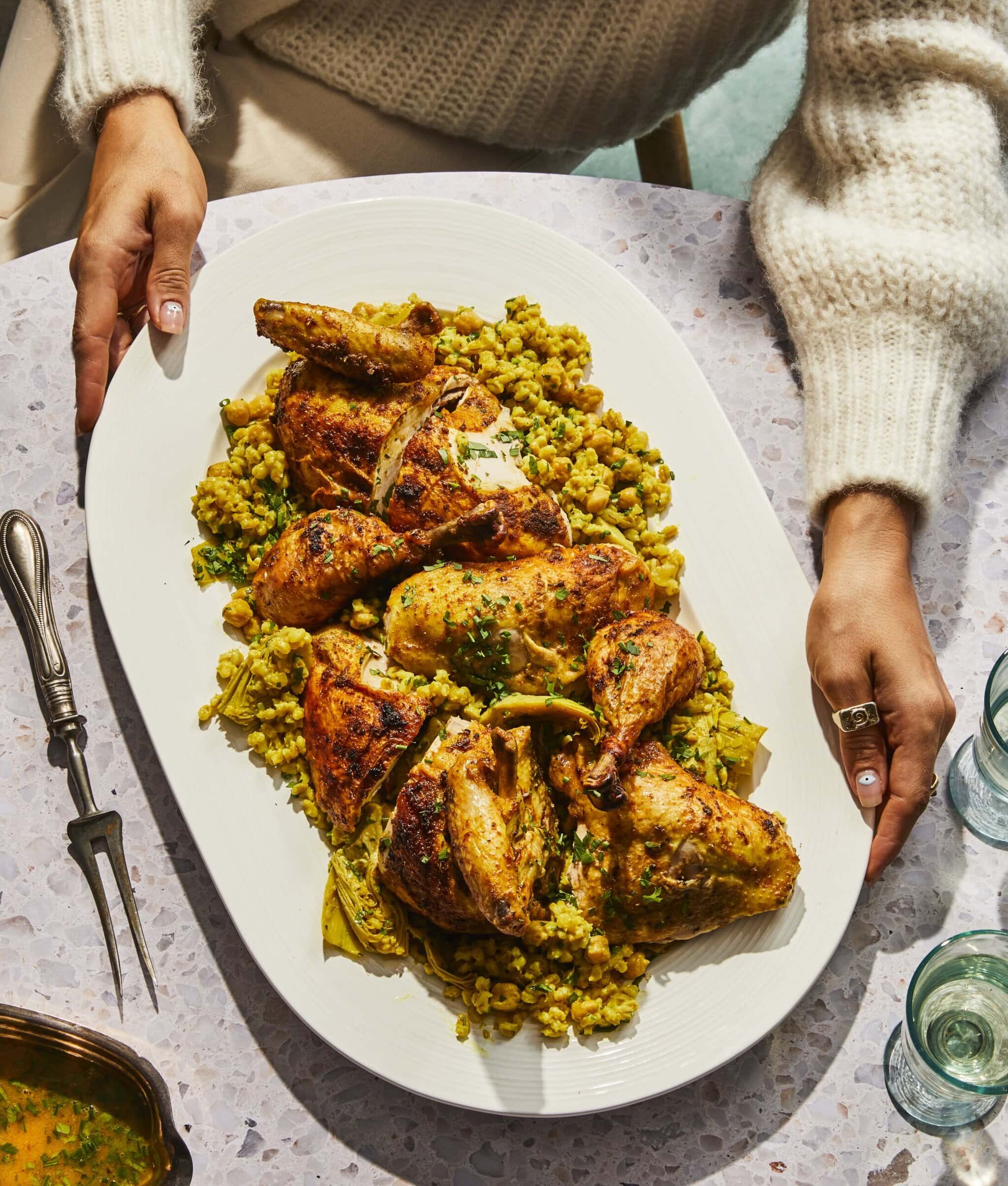 A person in a cream sweater holds a large white platter with roasted chicken pieces and golden couscous, garnished with herbs, on a speckled tabletop set for a meal.