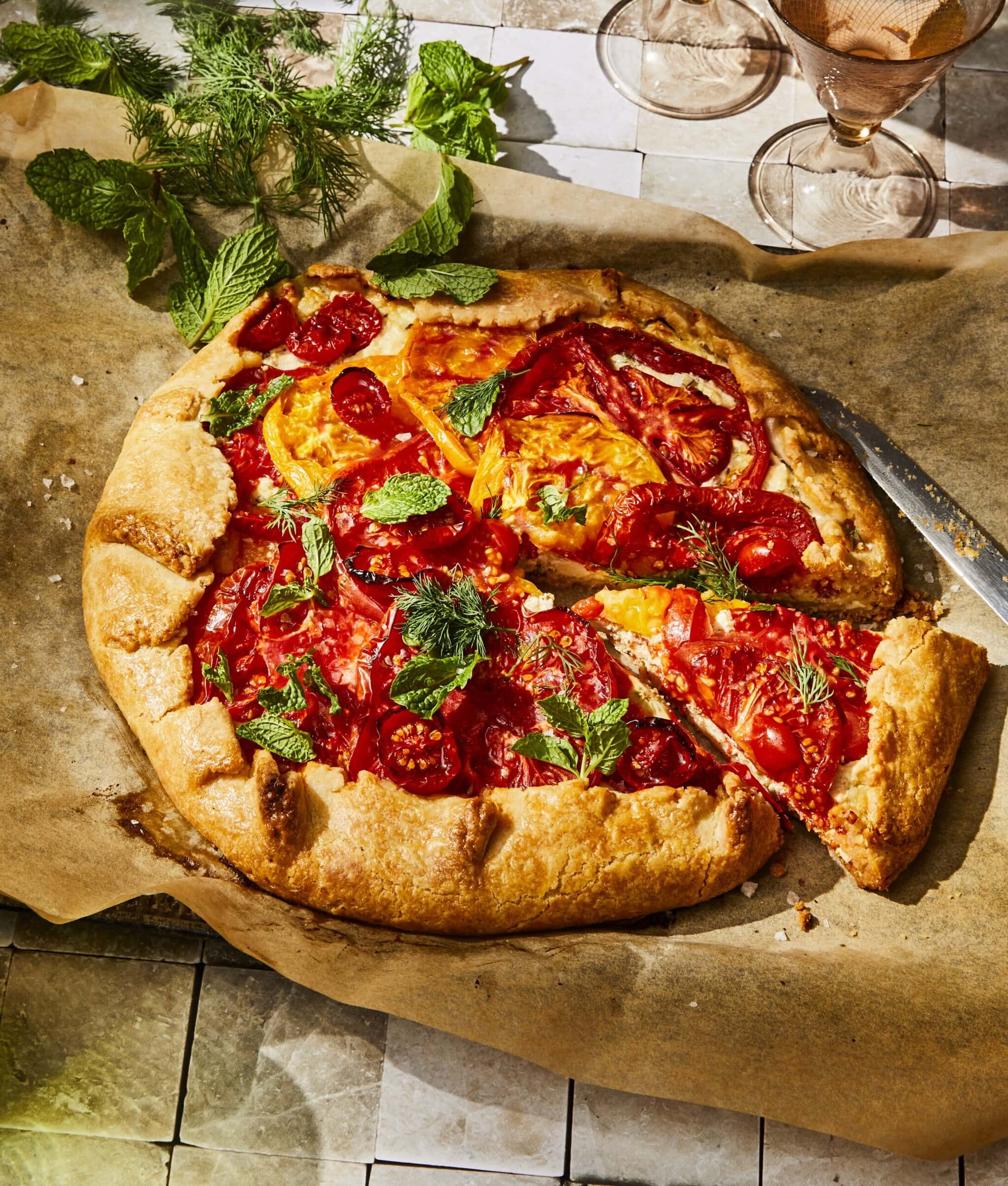 A rustic tomato galette sits on parchment paper, garnished with fresh herbs. A slice has been cut, revealing a flaky crust and vibrant red and yellow tomato filling. Nearby are herbs and empty glassware on a tiled surface.