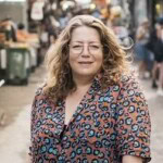Adeena Sussman with curly hair and glasses, wearing a patterned shirt, stands outdoors in a busy marketplace with blurred people and stalls in the background.