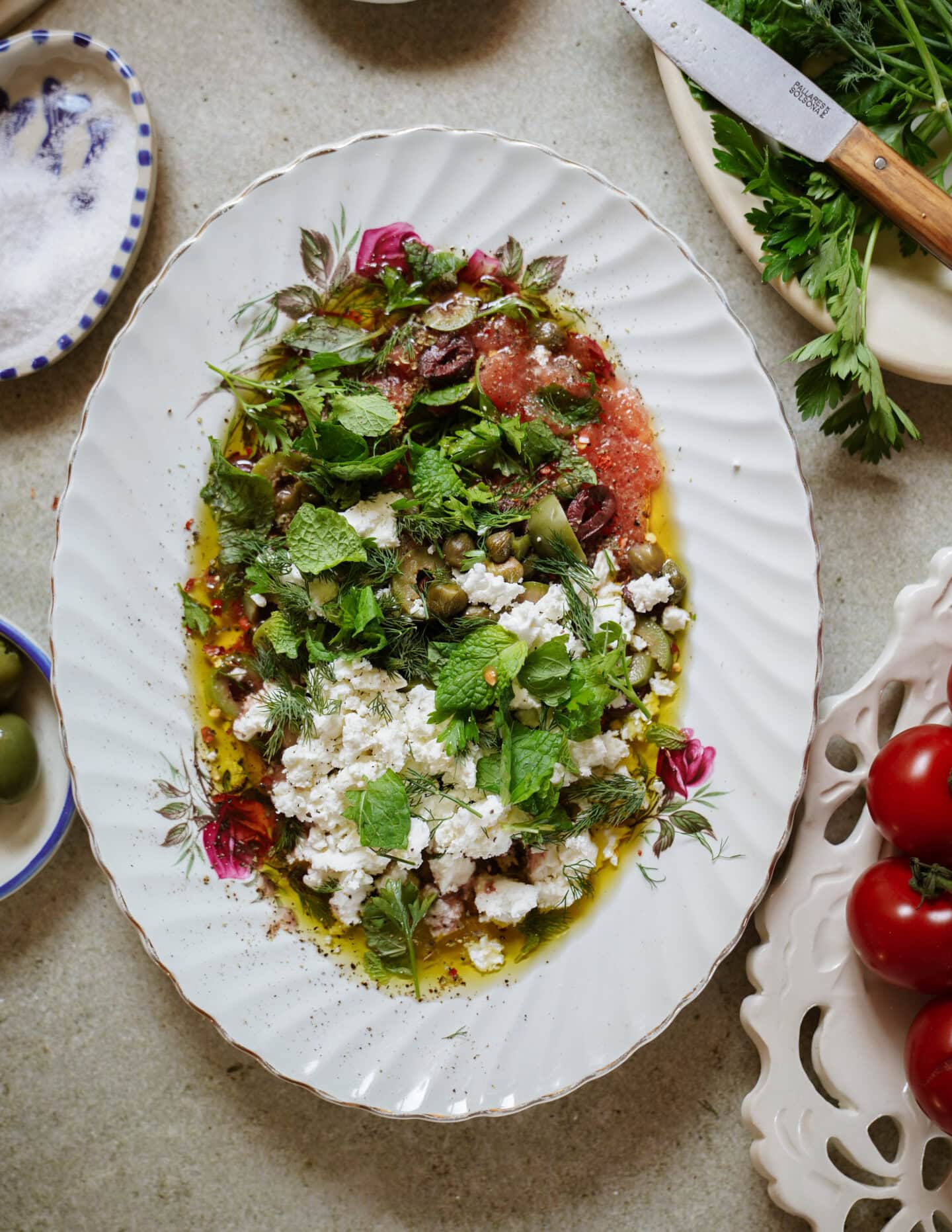 An oval platter with a Mediterranean salad of crumbled feta, olives, chopped herbs, olive oil, and tomatoes, garnished with fresh mint. Surrounding the platter are salt, tomatoes, herbs, and a knife.
