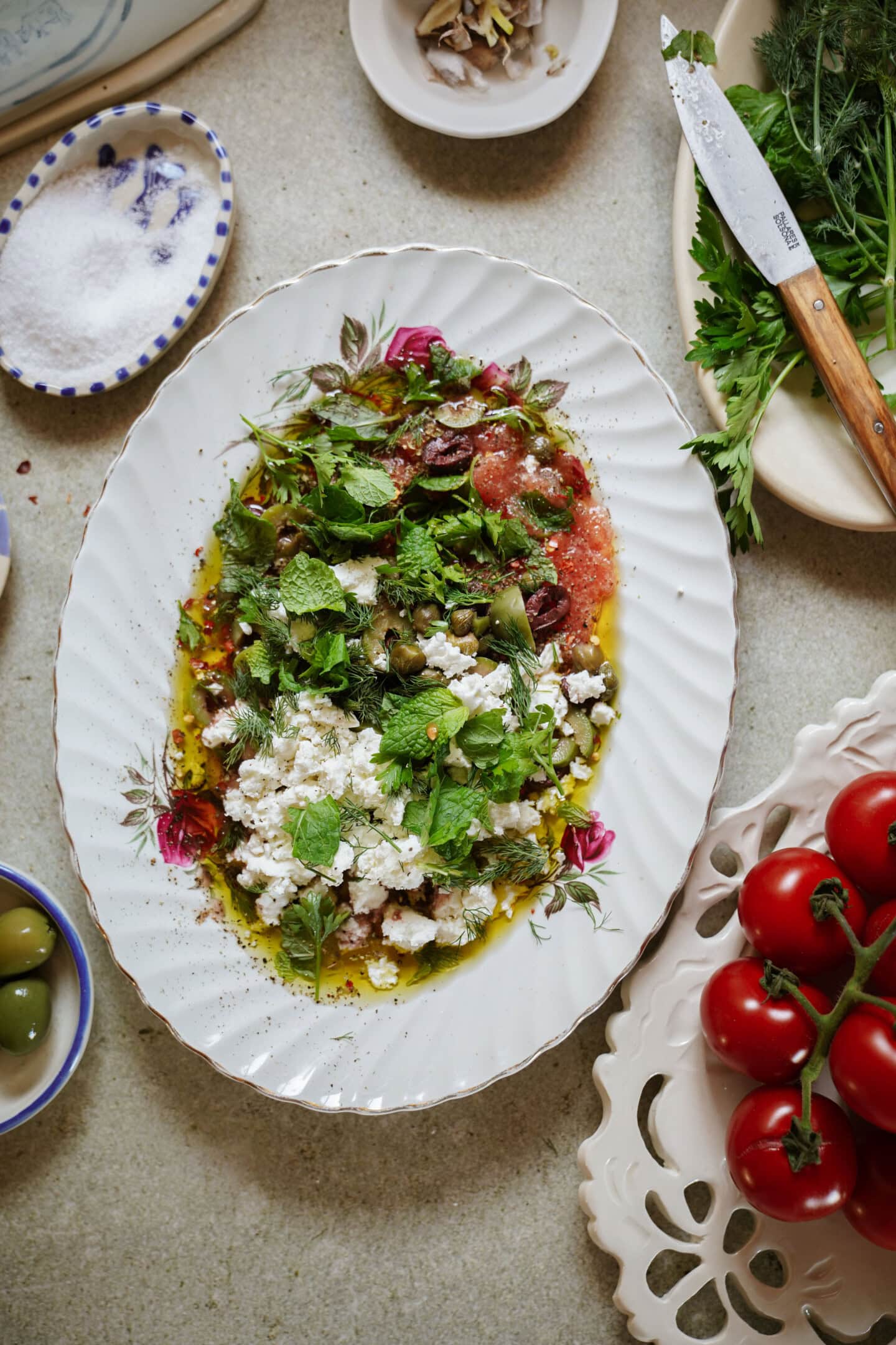 An oval plate with a Mediterranean salad featuring crumbled feta, fresh mint, herbs, olives, and tomatoes, drizzled with olive oil. Surrounding the plate are tomatoes, olives, herbs, salt, and a knife on a tabletop.