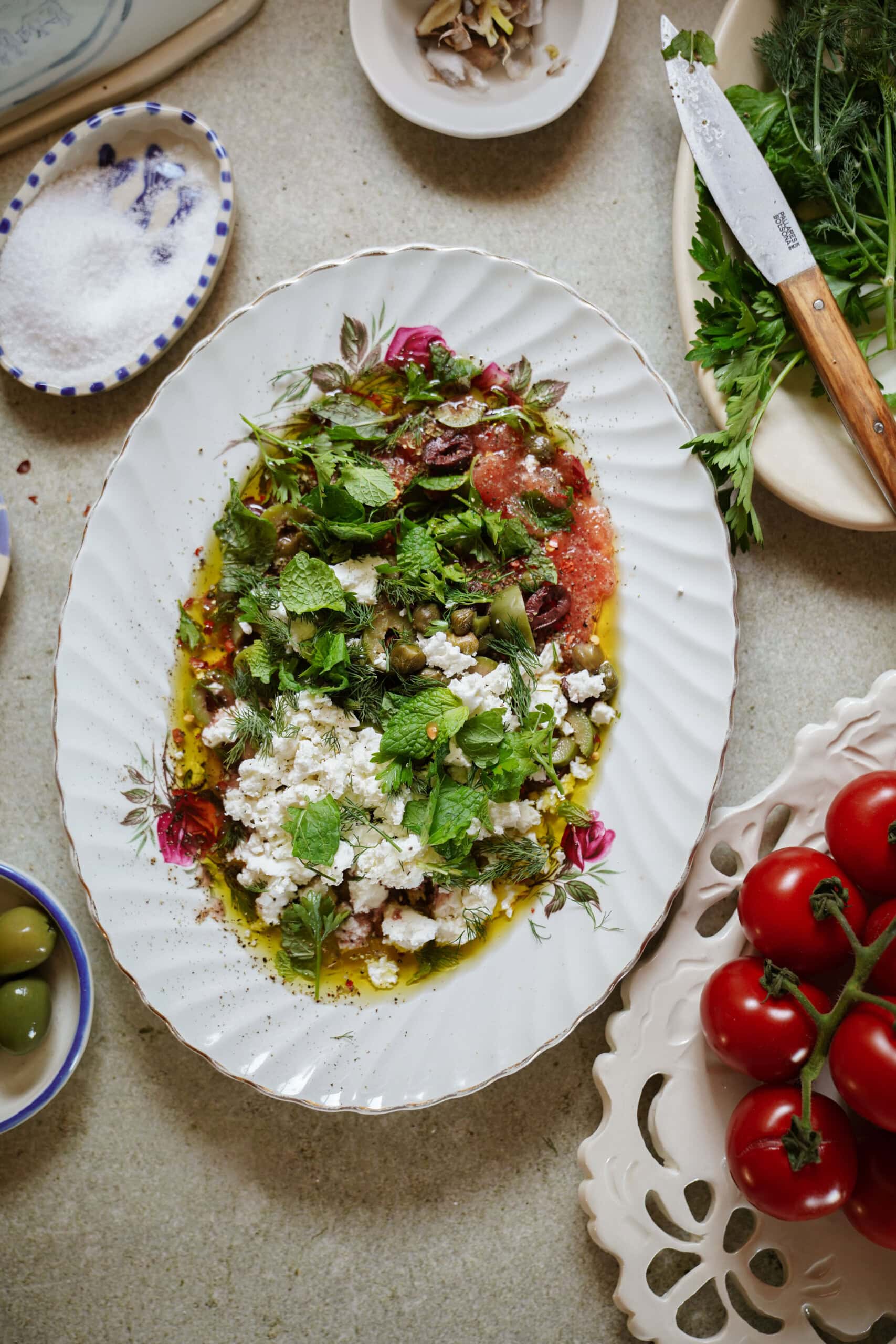 An oval plate with a Mediterranean salad featuring crumbled feta, fresh mint, herbs, olives, and tomatoes, drizzled with olive oil. Surrounding the plate are tomatoes, olives, herbs, salt, and a knife on a tabletop.