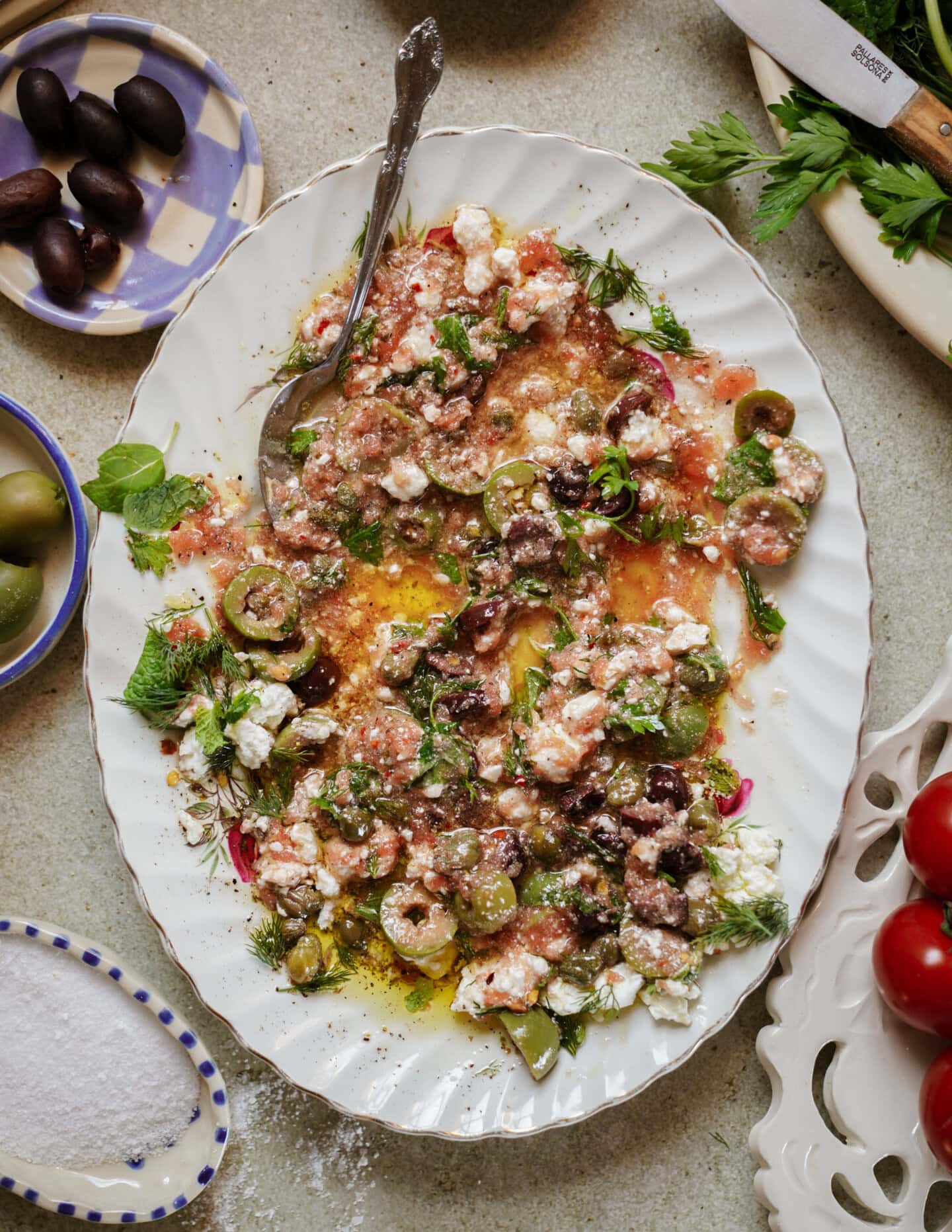 A large oval platter holds a rustic salad with crumbled feta, sliced green and black olives, chopped tomatoes, herbs, and olive oil. A spoon rests on the plate. Dishes of olives, salt, and vegetables surround the platter.