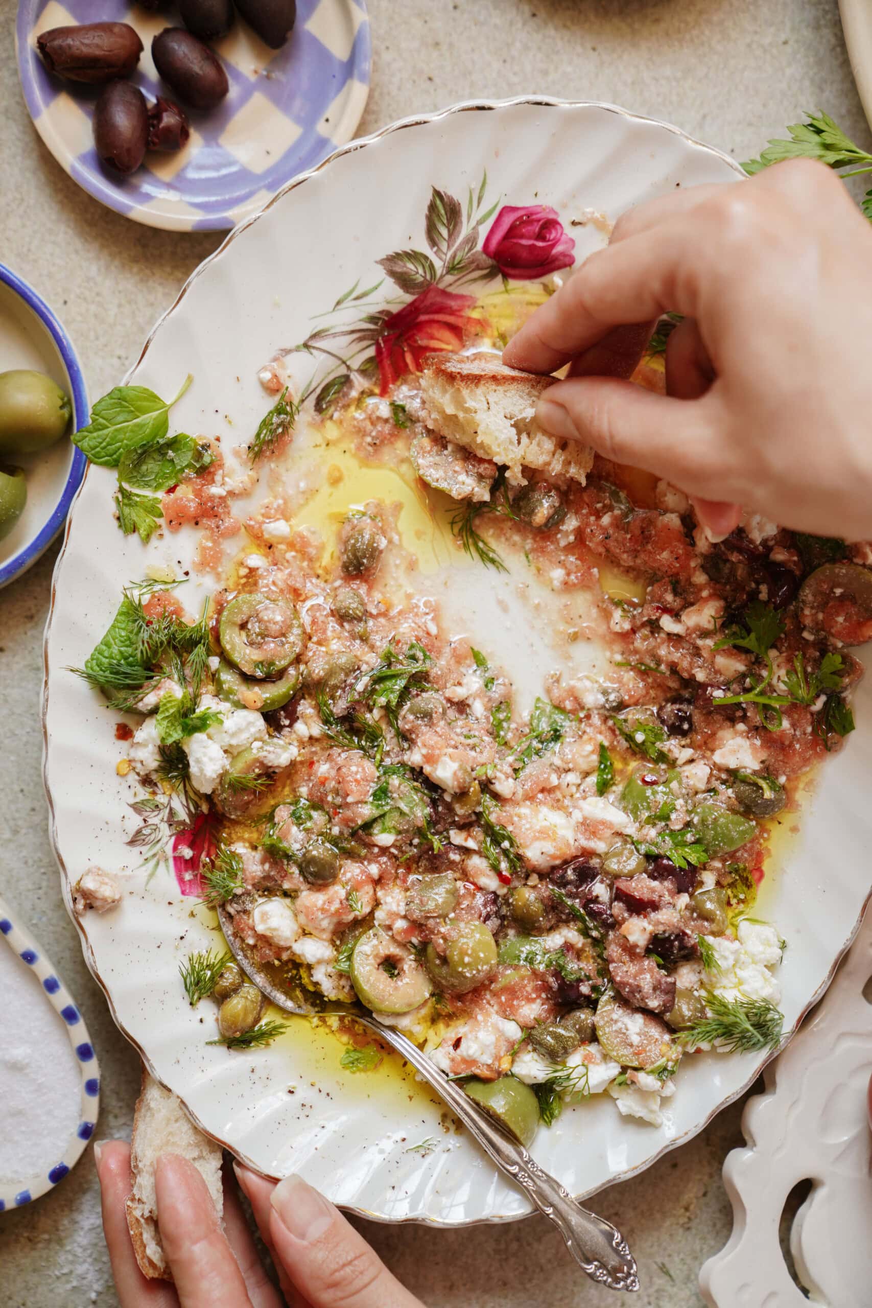 A hand dips bread into a platter of olive oil, feta cheese, chopped olives, herbs, and tomatoes on a floral dish. Plates of olives and bread are visible nearby on a light tabletop.