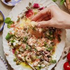 A hand dips bread into a colorful, partially eaten Mediterranean salad with feta, olives, herbs, and olive oil on a floral plate. Other food items are visible around the plate.
