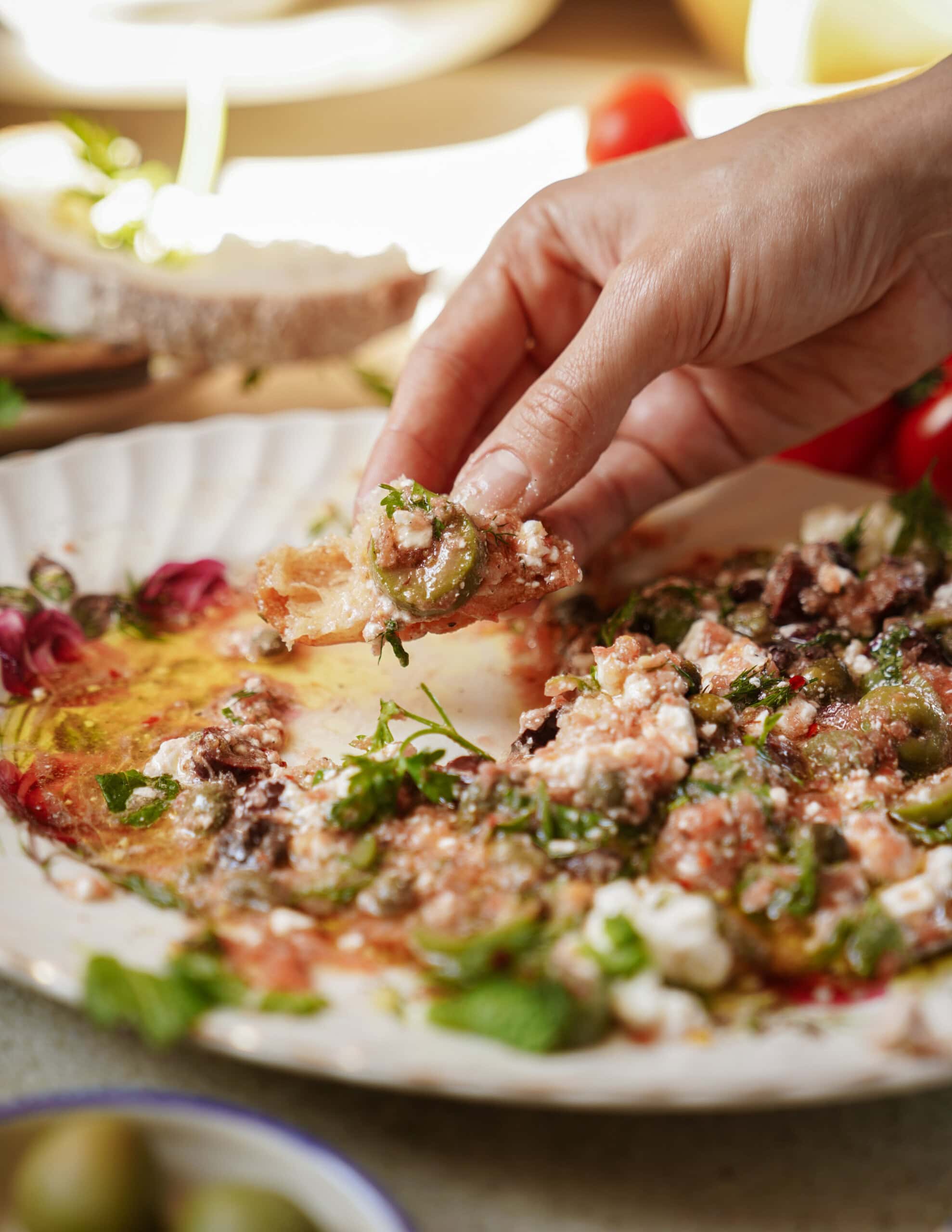 A hand scoops a piece of bread through a plate of Mediterranean dip made with feta cheese, olives, herbs, and olive oil, with fresh ingredients visible in the background.