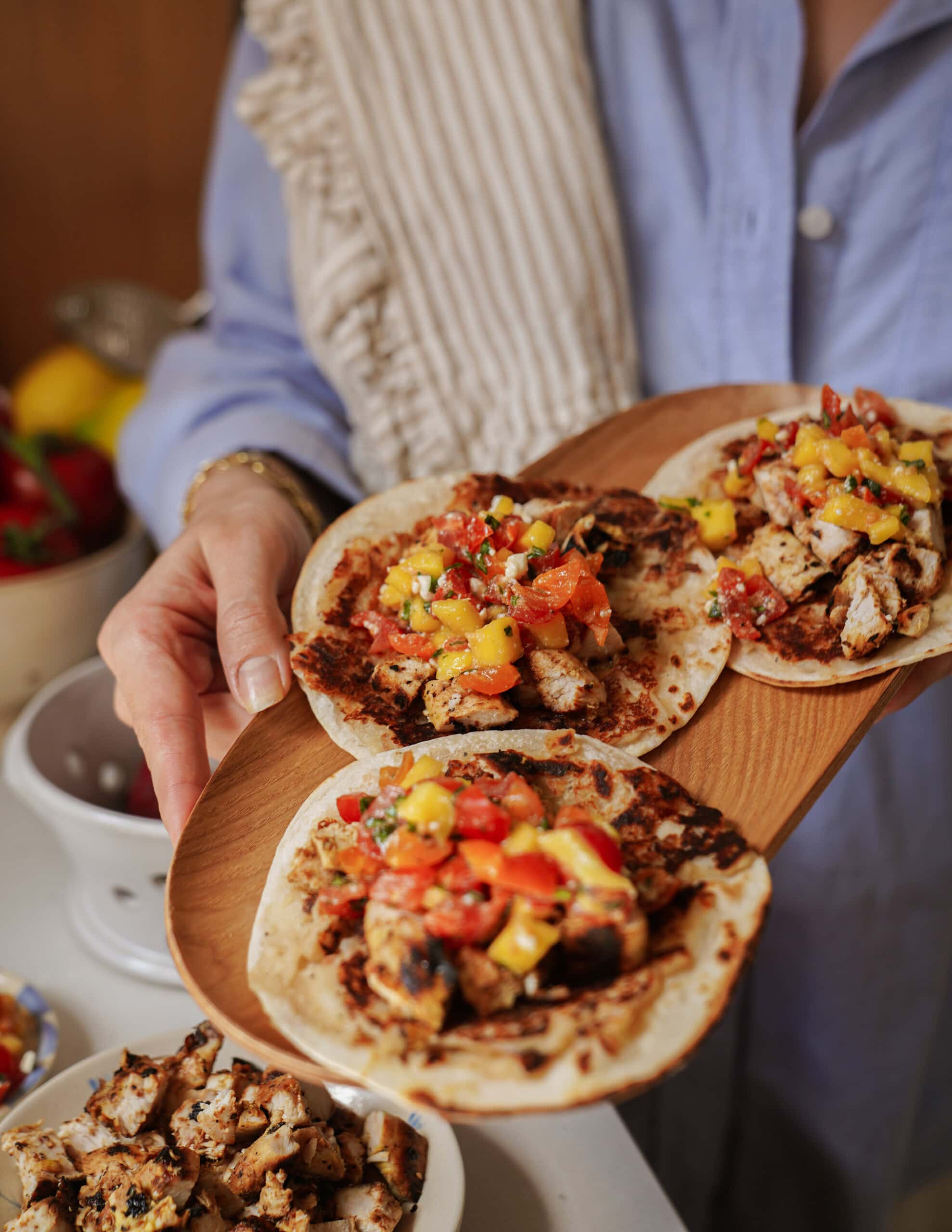 A person in a blue shirt and striped towel holds a wooden platter with three tacos topped with grilled chicken and colorful salsa. A bowl of extra chicken and fresh produce are visible on the counter nearby.
