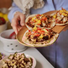 A person holds a wooden plate with three tacos filled with grilled chicken and colorful salsa. A bowl of diced chicken and kitchen items are visible on the table in the background.