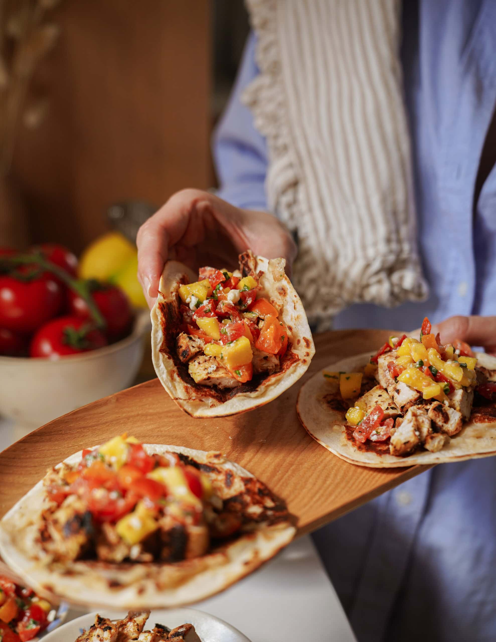 A person wearing a blue shirt and striped towel holds a wooden platter with tacos filled with grilled chicken, tomatoes, and mango salsa. A bowl of tomatoes and lemons is blurred in the background.
