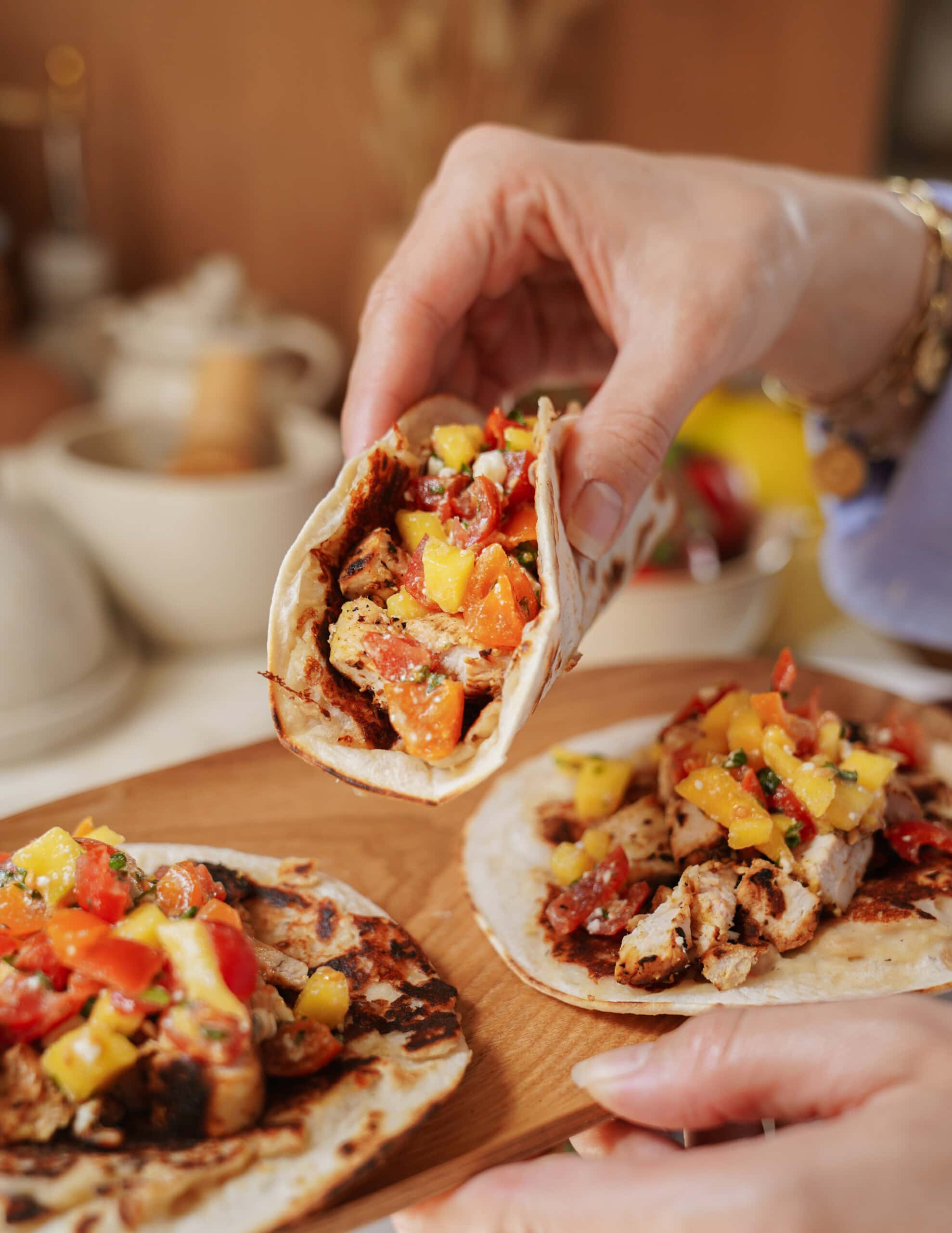 A hand holds a taco filled with grilled chicken, diced tomatoes, and mango salsa above a wooden board with two more loaded tacos. The scene is bright and inviting, highlighting the colorful, fresh ingredients.