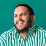 Dan Pelosi with a beard and short hair smiles while looking upward. He is wearing a green and white vertically striped shirt and is posed in front of a solid teal background.