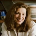 Diane Kochilas with long brown hair wearing a white blouse smiles warmly at the camera, with shelves of plates and a blue teapot visible in the background.