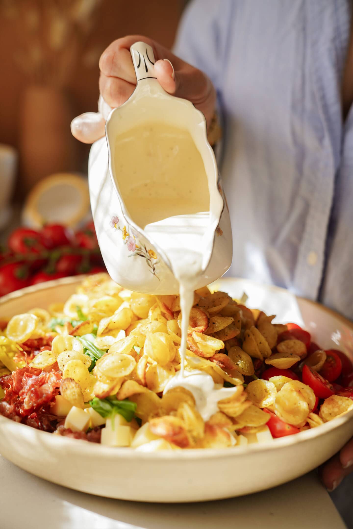 A person pours creamy white sauce from a floral pitcher onto a dish of sliced cooked potatoes, tomatoes, and herbs in a ceramic bowl. The background is softly blurred, highlighting the food.