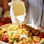 A person pours creamy white sauce from a decorative jug over a bowl filled with orecchiette pasta, diced vegetables, cheese cubes, and sundried tomatoes. The food looks fresh and colorful.