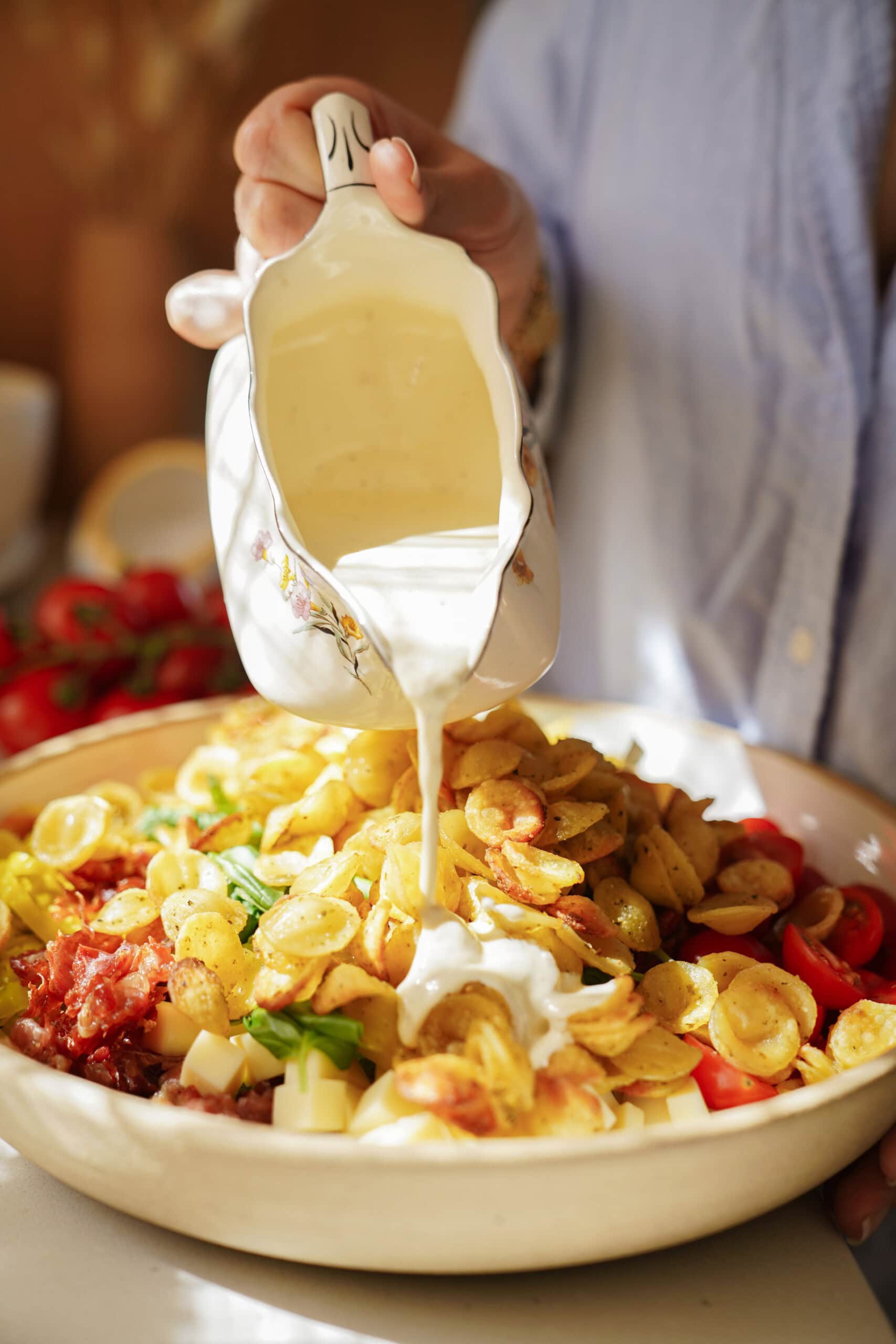 A person pours creamy white sauce from a decorative jug over a bowl filled with orecchiette pasta, diced vegetables, cheese cubes, and sundried tomatoes. The food looks fresh and colorful.