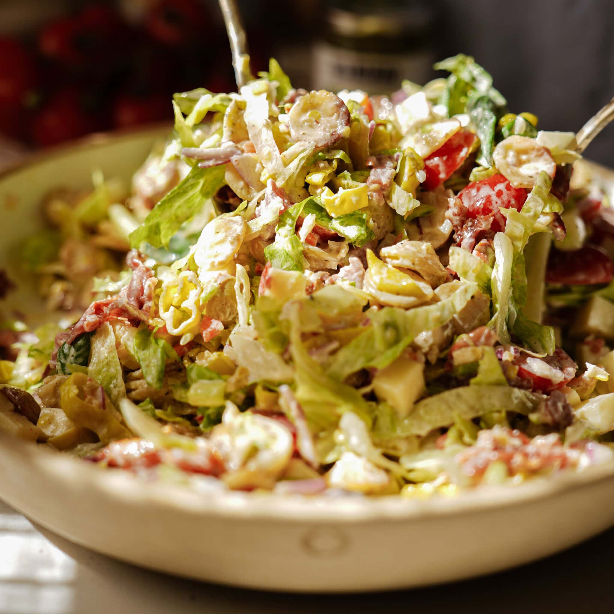 A close-up of a large bowl filled with a colorful chopped salad, featuring lettuce, tomatoes, cheese, and various vegetables, all mixed with a creamy dressing. Two serving utensils are visible in the bowl.